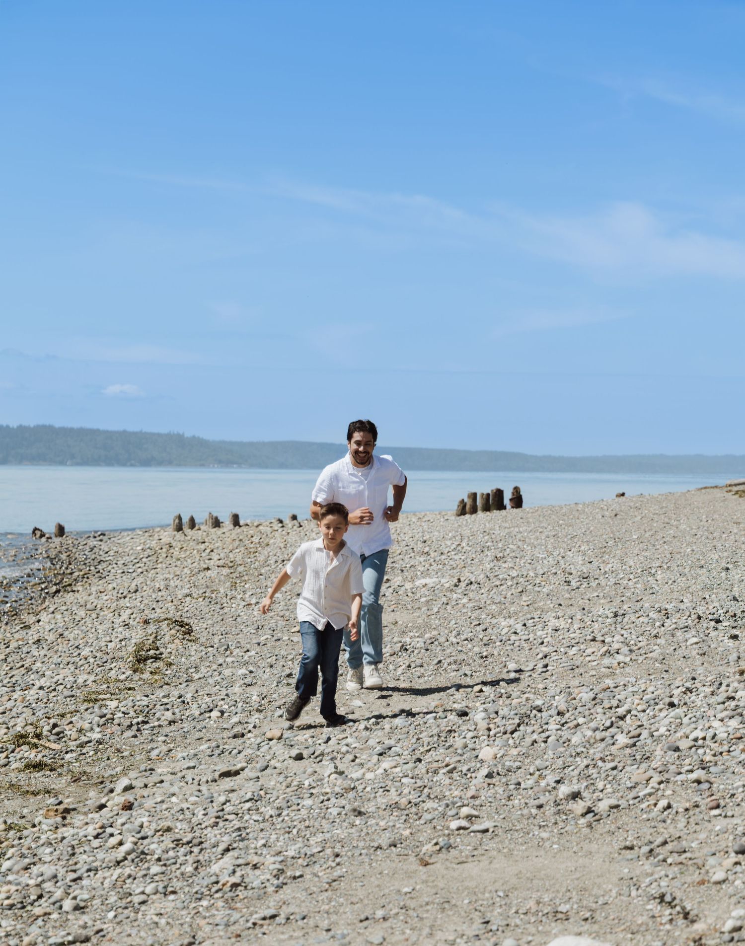 Father and young son run together along a sunny rocky beach beside calm blue water.