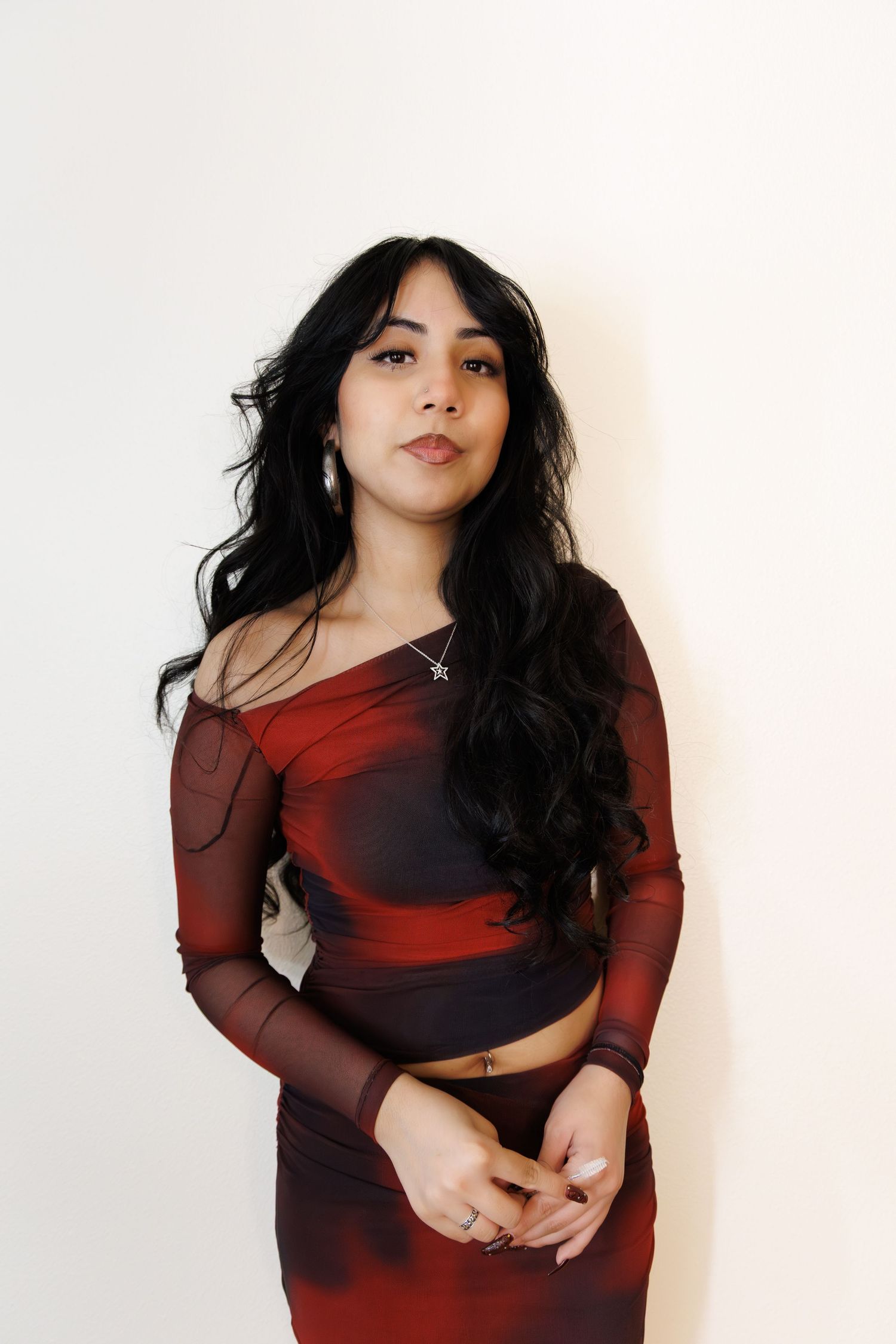 Young woman with long black hair wearing a red mesh crop top and skirt set, posing against a white wall.