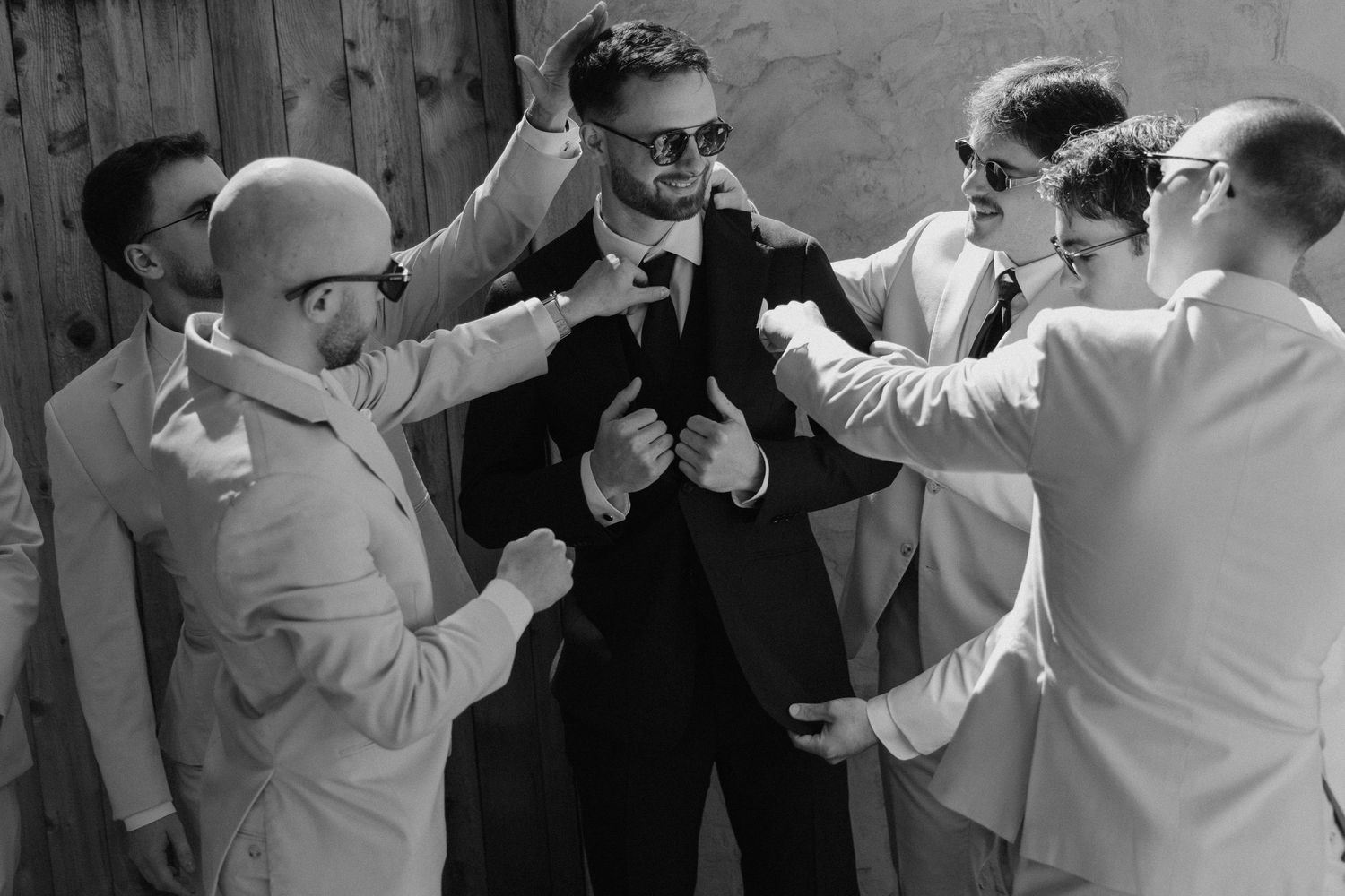 Groom in black suit surrounded by groomsmen in white suits adjusting his tie in black and white photo.