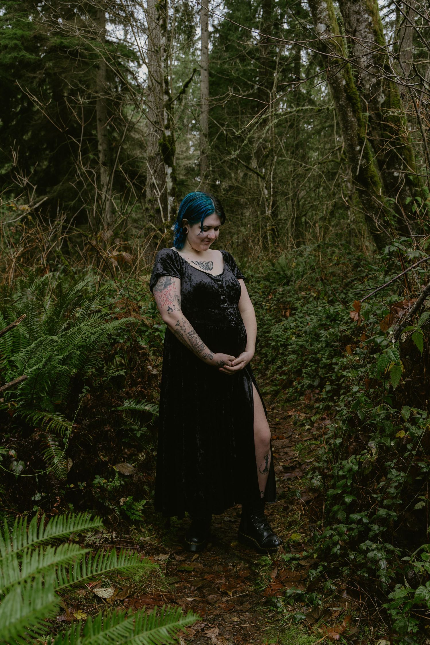 Woman with blue hair and tattoos in black dress standing on a forest path surrounded by ferns.