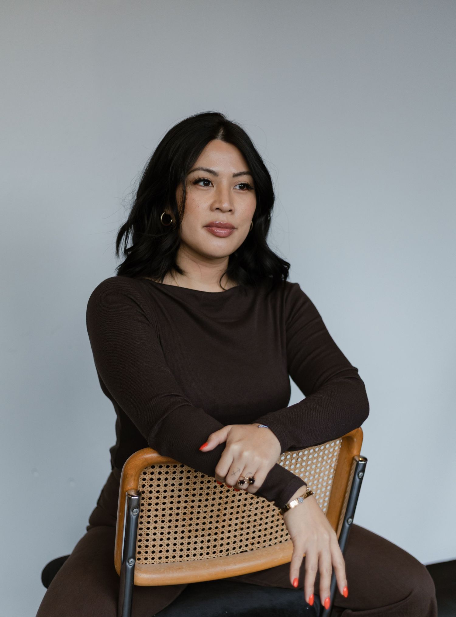 Woman in brown long-sleeve top seated on a wicker chair posing confidently against a light gray background.