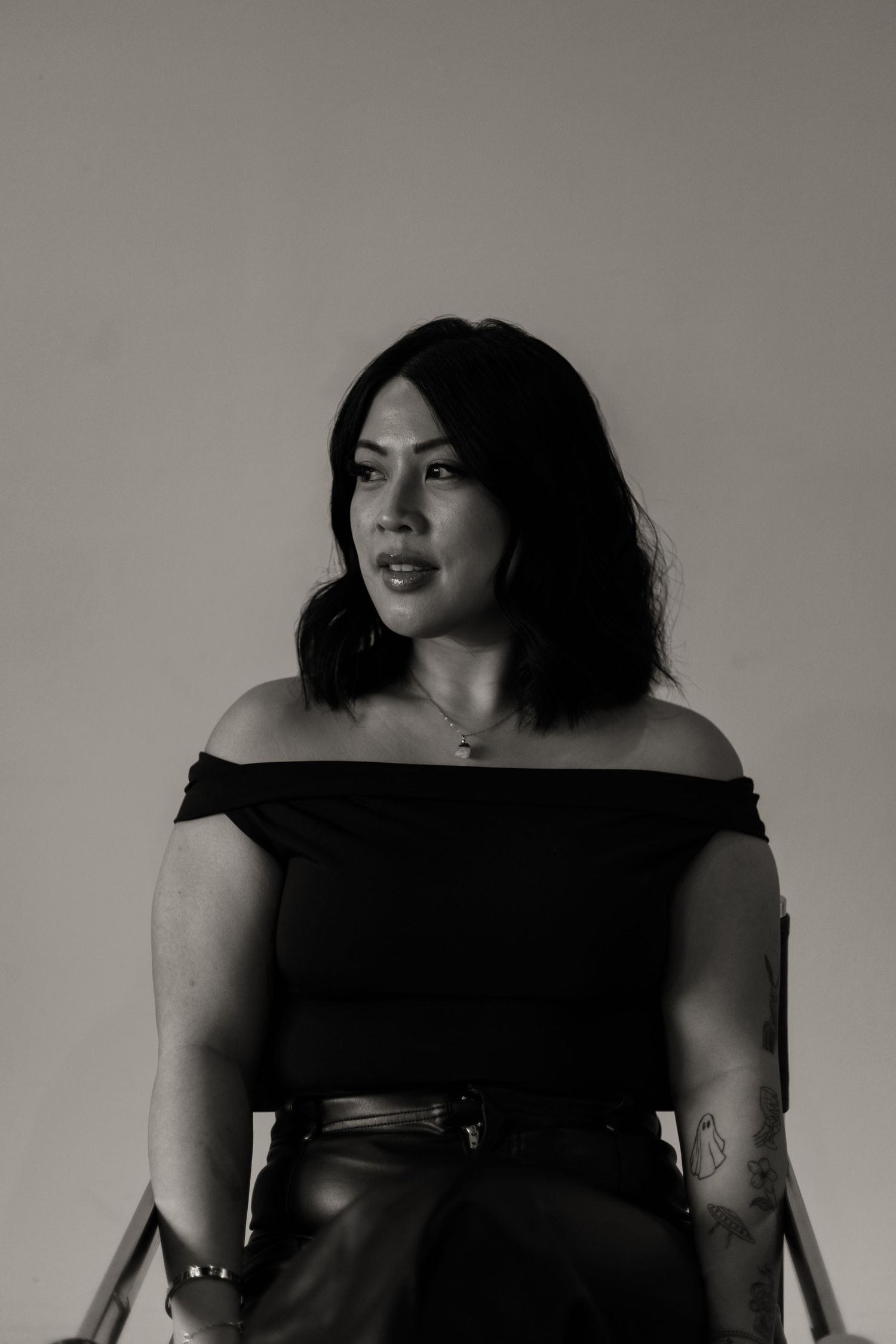 Black and white portrait of a woman in off-shoulder top seated in a chair against neutral background.