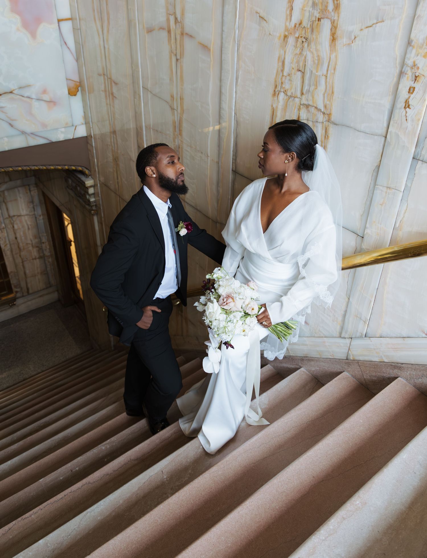Black bride and groom sharing a romantic moment on marble staircase, bride holding white floral bouquet.
