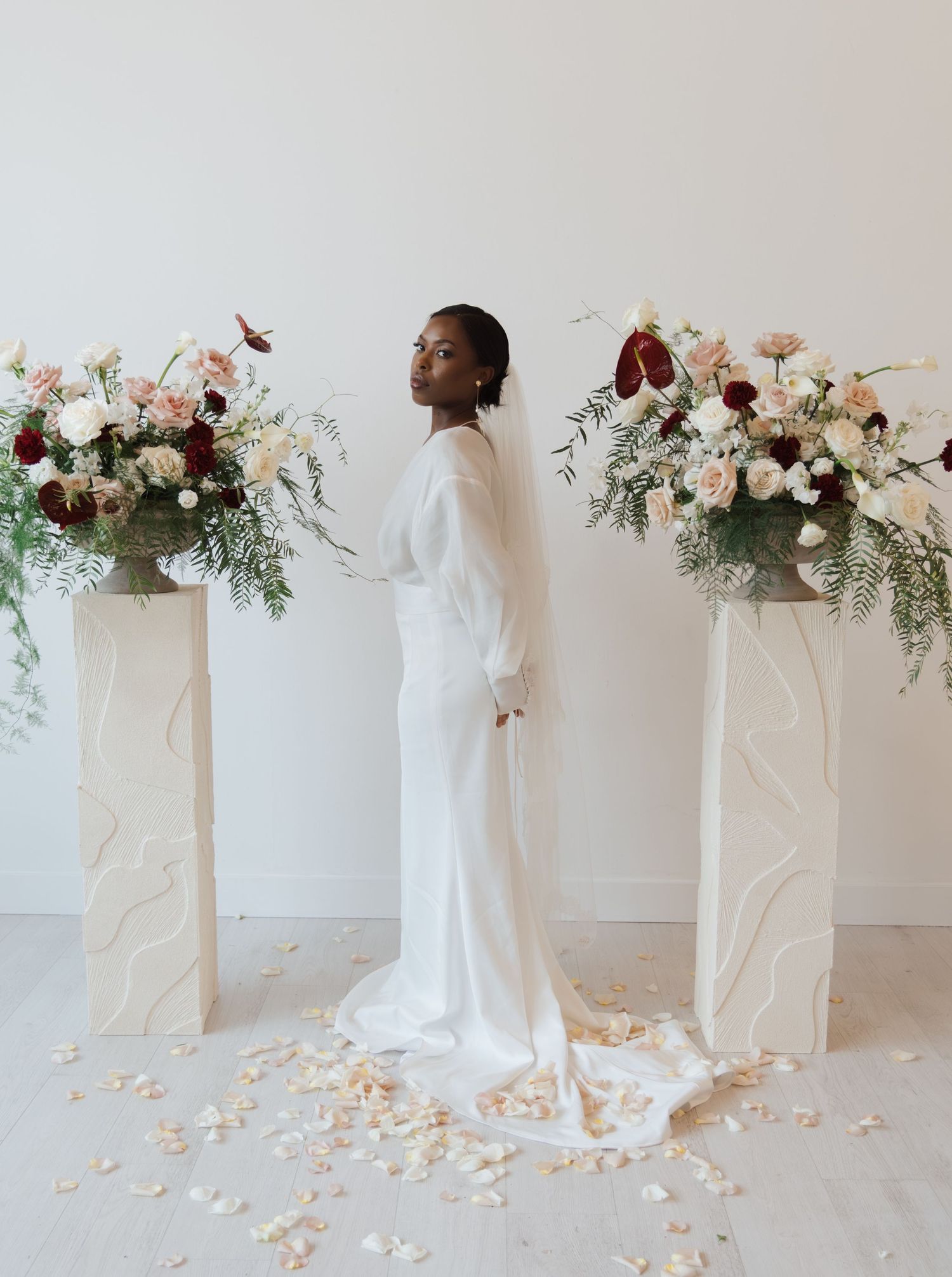 Bride in white long-sleeve gown standing between floral arrangements with scattered rose petals on floor.