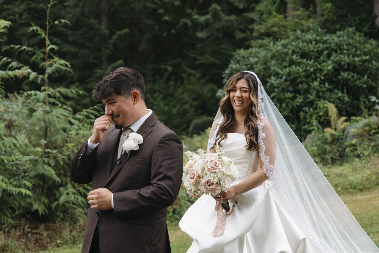 Emotional groom wipes tears during first look with bride holding bouquet in lush forest wedding setting.