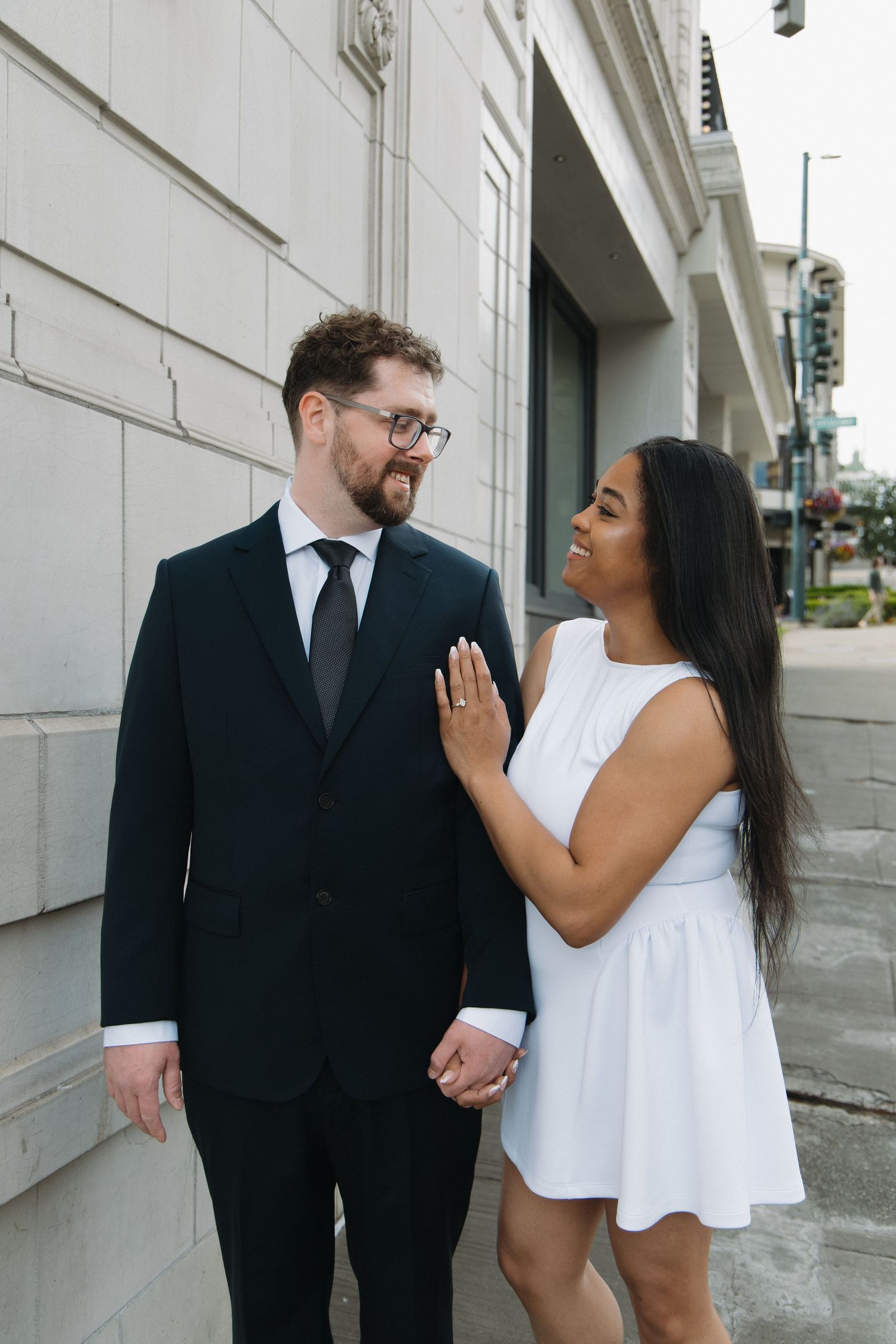Interracial couple sharing a tender moment outdoors, woman in white dress placing hand on mans chest while smiling.