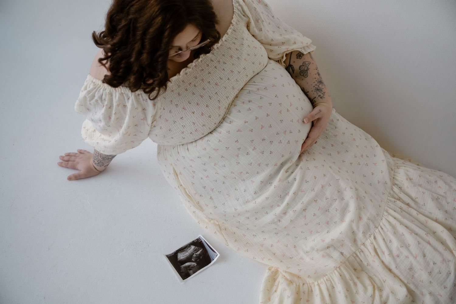 Overhead view of pregnant woman in cream floral dress holding ultrasound photo on white floor.