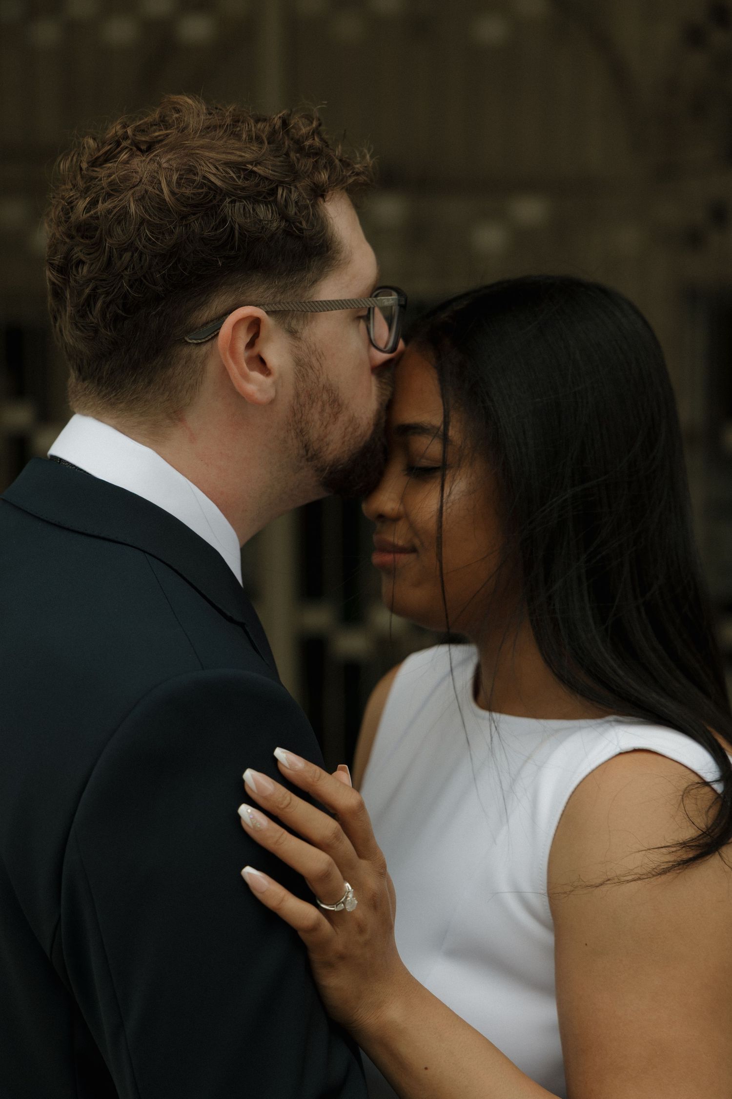 A groom in a dark suit kisses his bride on the forehead as she closes her eyes wearing a white dress.