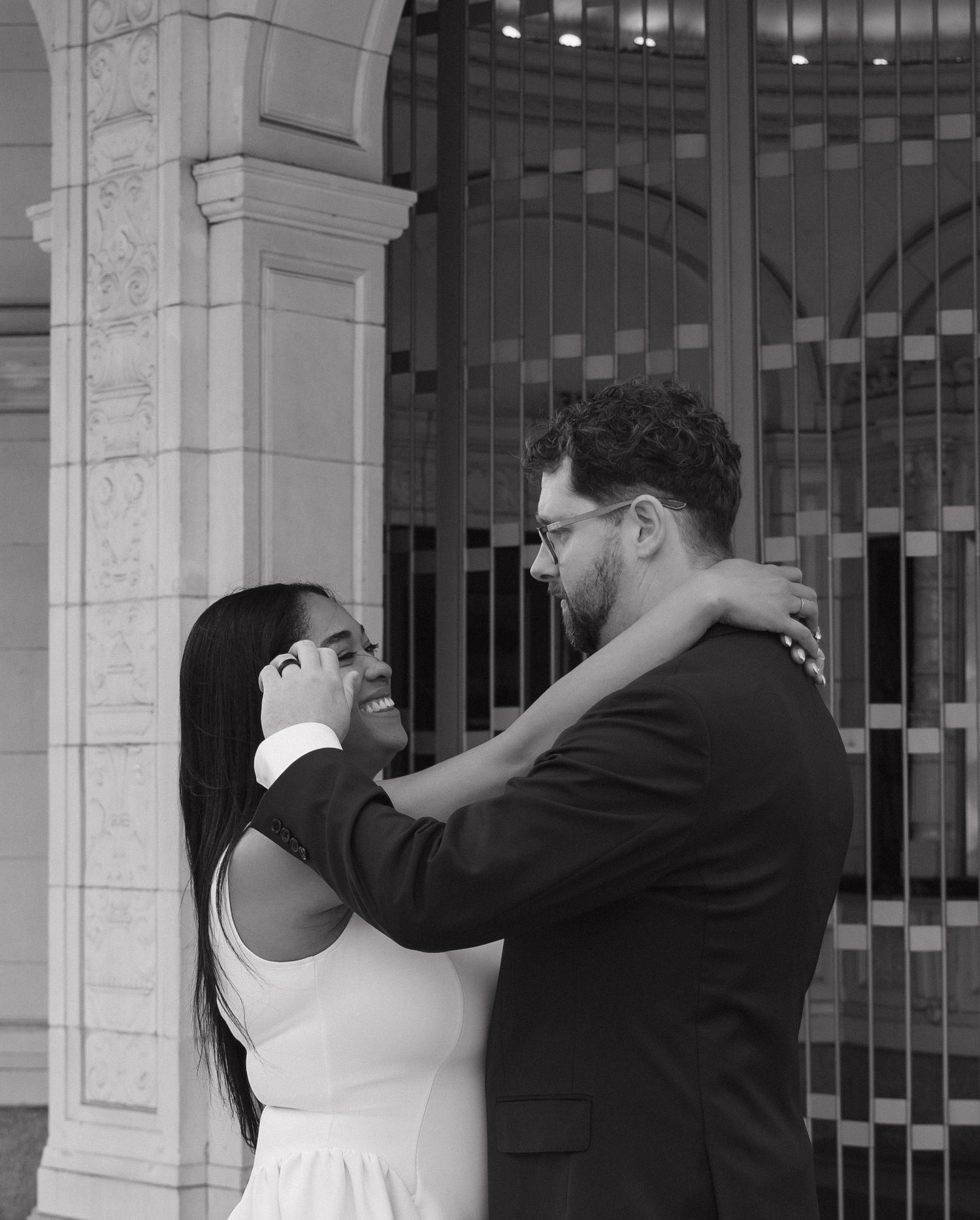 Black and white photo of a smiling couple embracing near an elegant arched building entrance.