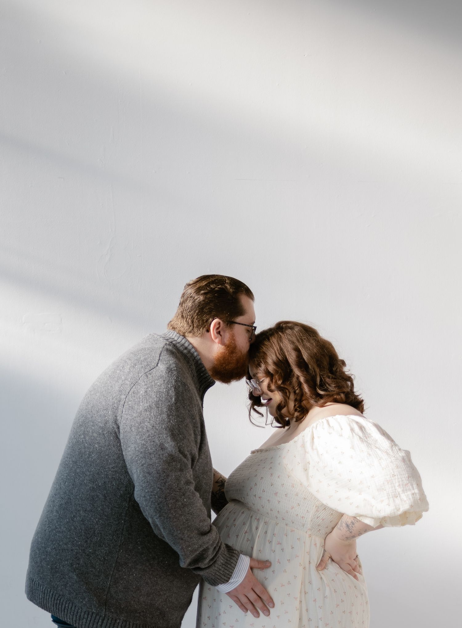 Couple sharing a tender forehead kiss while cradling pregnant belly in soft natural light.