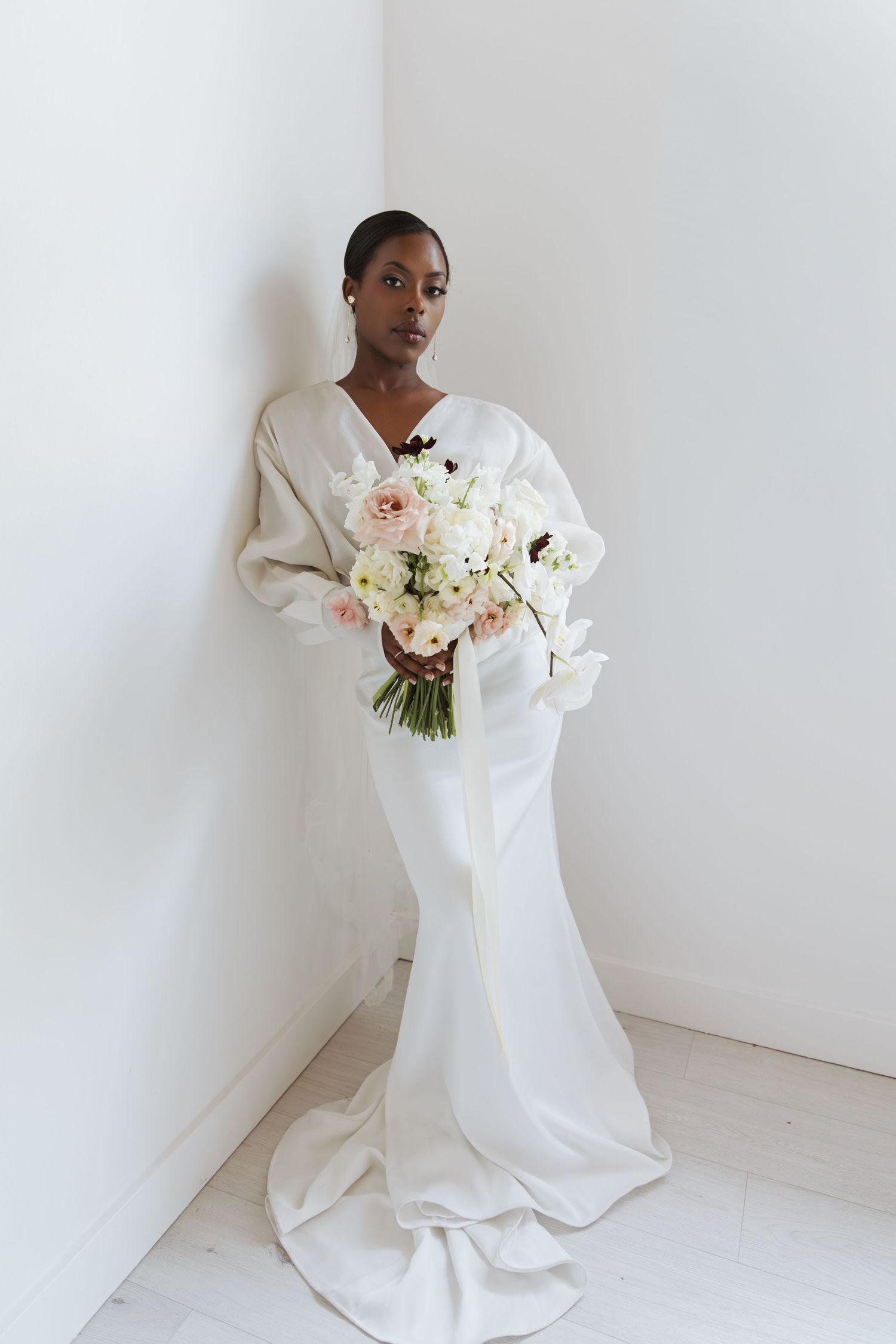 Elegant Black bride in white mermaid gown with oversized sleeves holding a pastel bouquet against white wall.