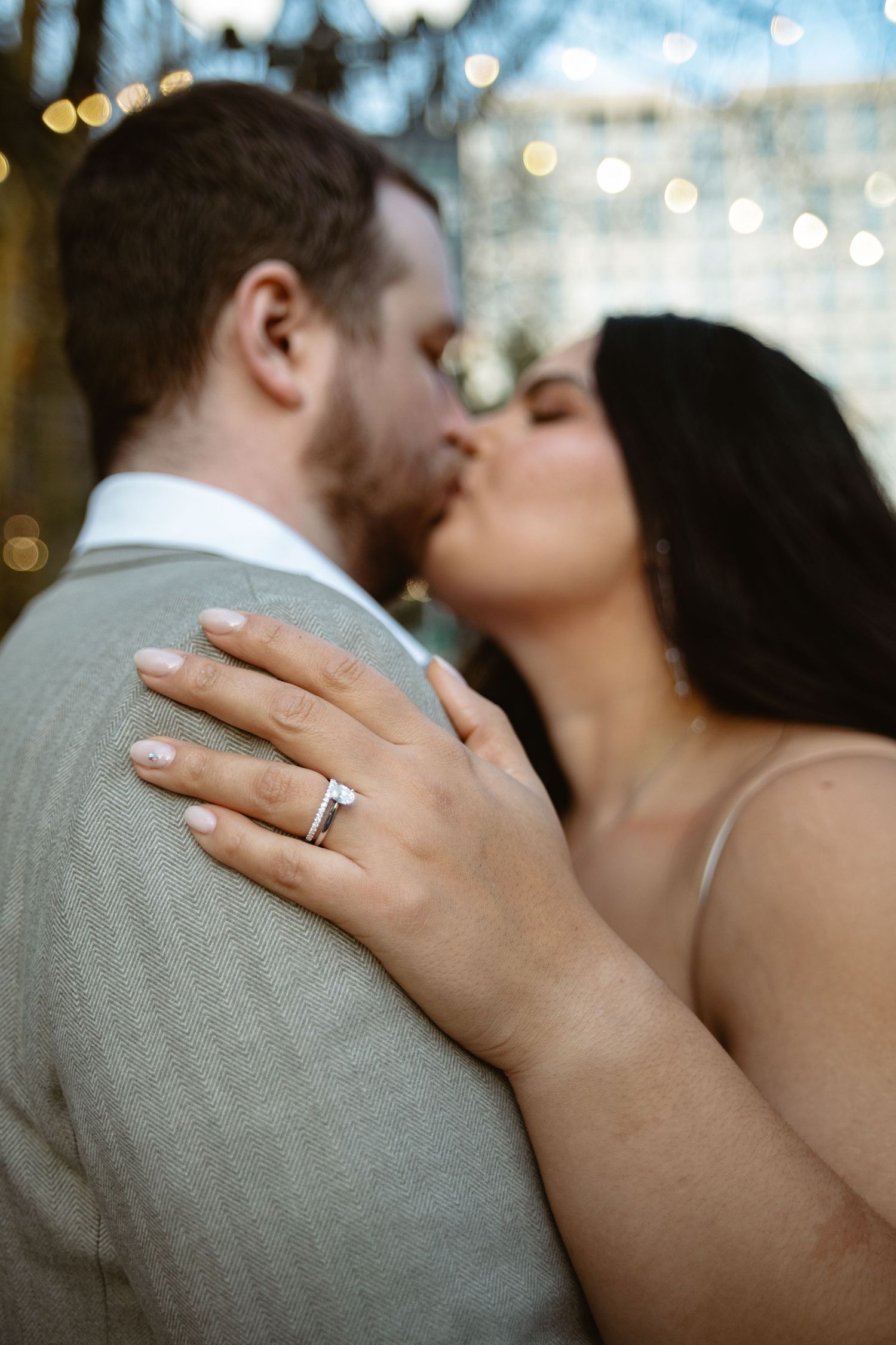 Close-up of bride kissing groom outdoors with diamond engagement ring prominently displayed.