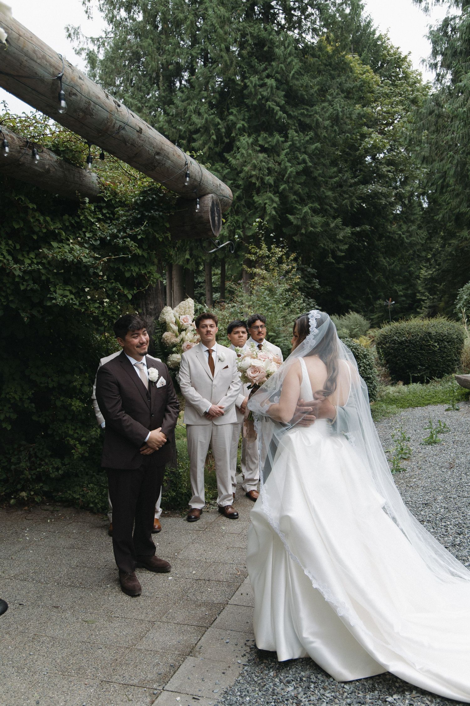 Groom in brown suit smiles as bride in white gown approaches outdoor wedding altar with groomsmen.