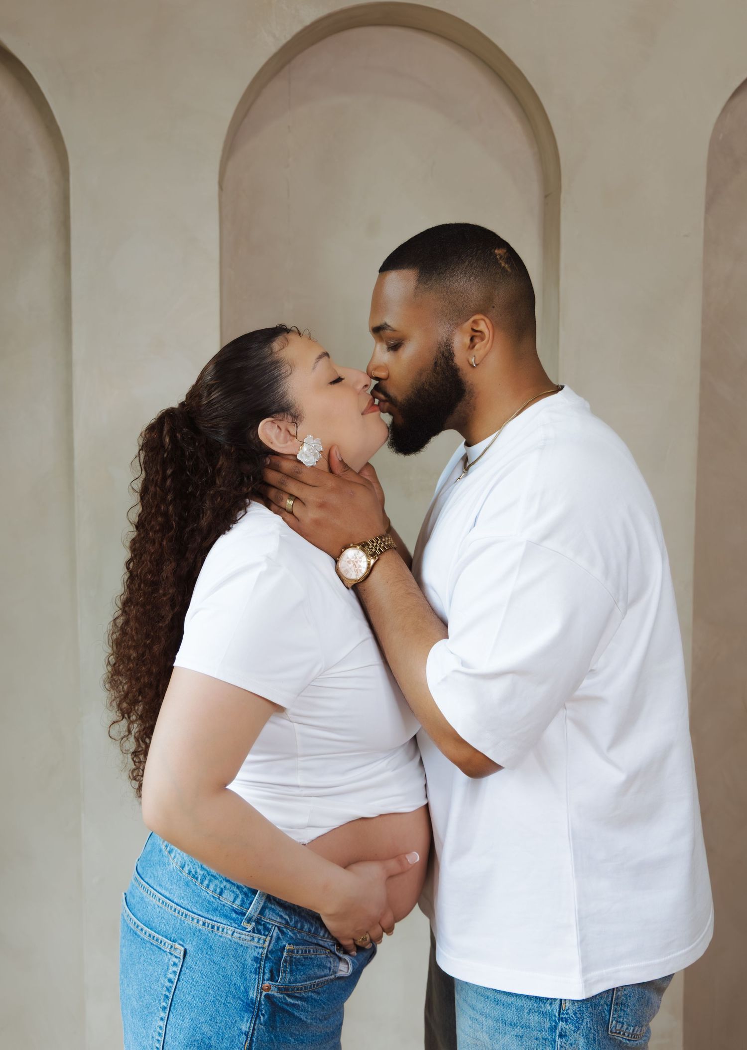 Pregnant couple in white outfits sharing a kiss while cradling baby bump against arched wall backdrop.