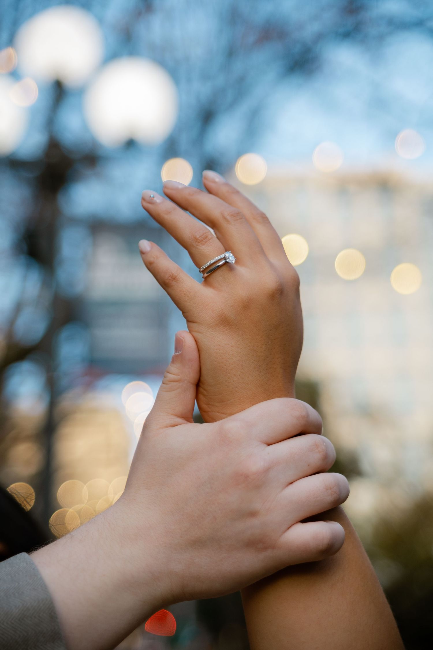 Close-up of hands displaying diamond engagement ring with bokeh lights in background.