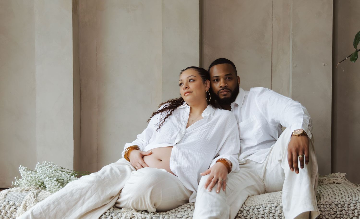 Expecting couple in matching white outfits sitting together relaxed against a neutral textured wall.
