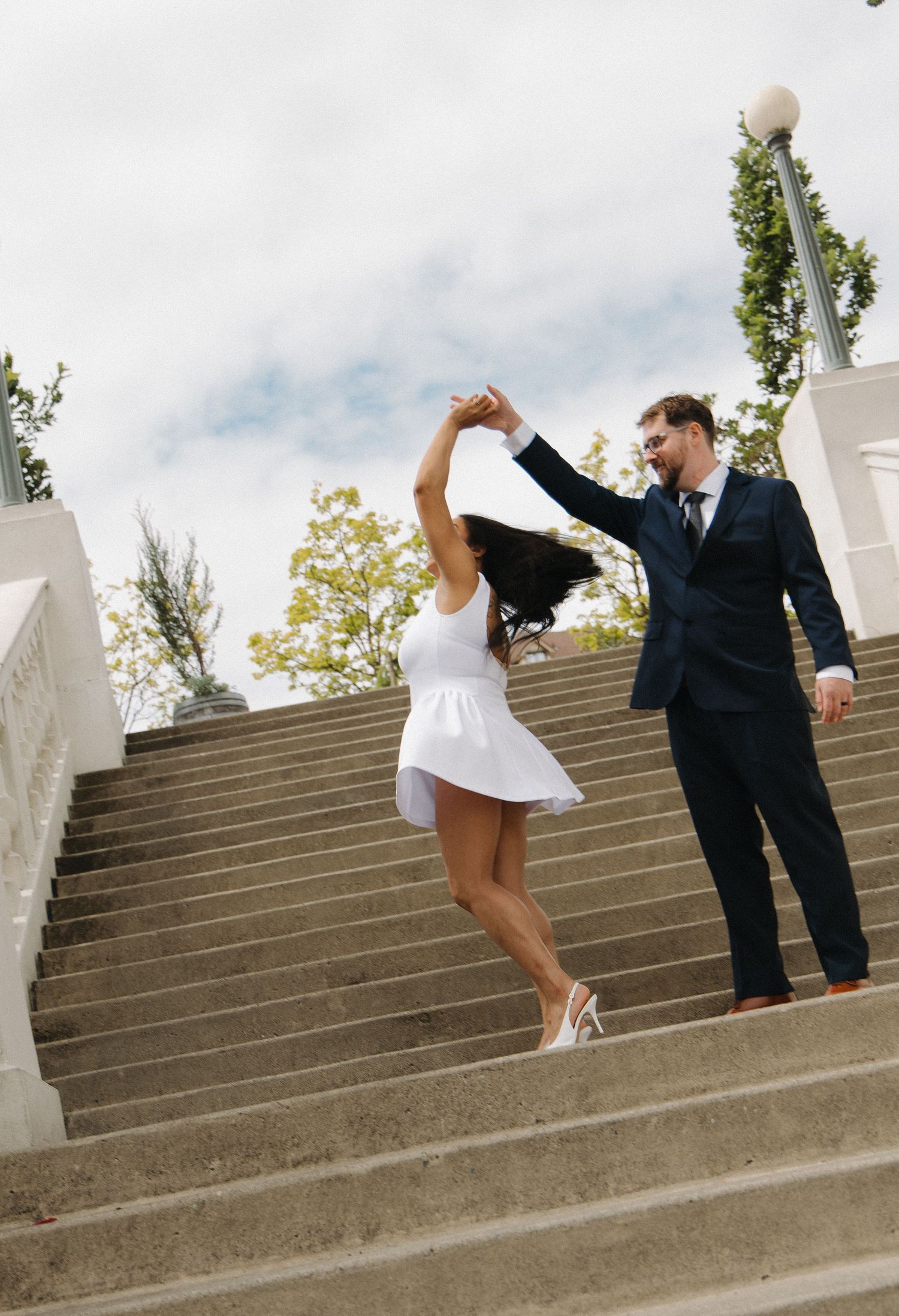 Groom spinning bride in white dress on outdoor staircase during wedding photo session.