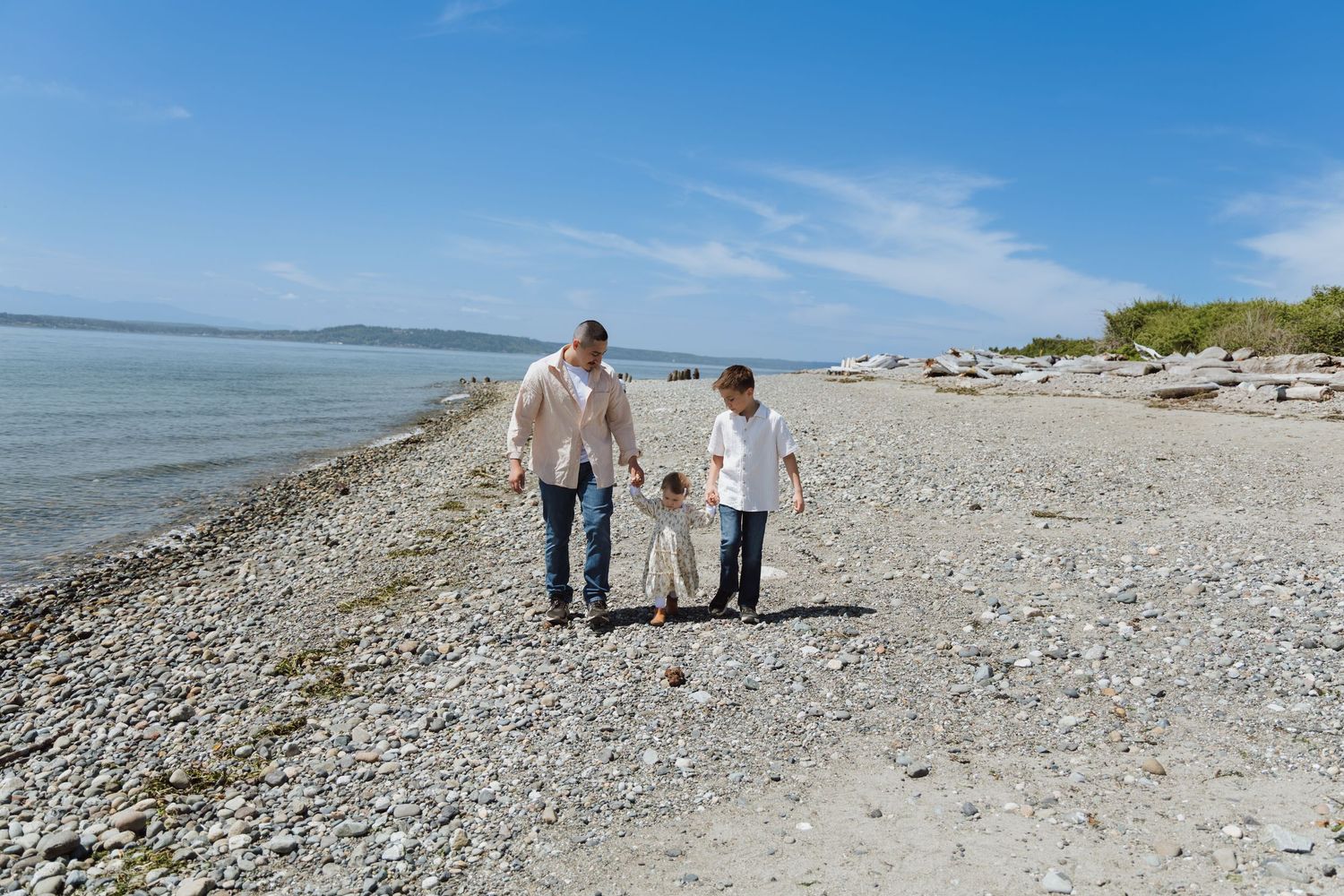 Family of three walking on a rocky beach on a sunny day with calm blue waters in the background.
