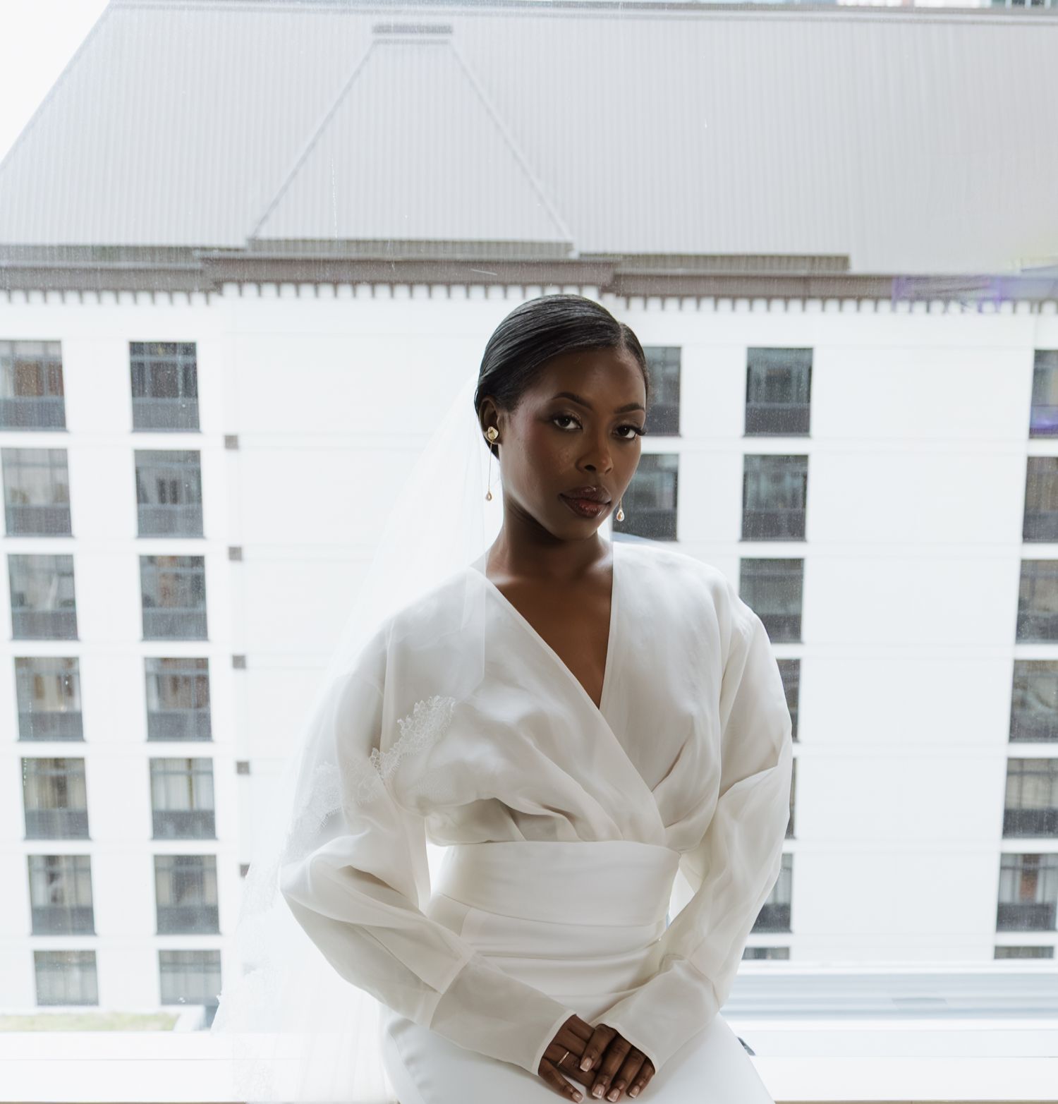 Elegant bride in white wrap gown with obi belt poses confidently by hotel window with city skyline backdrop.