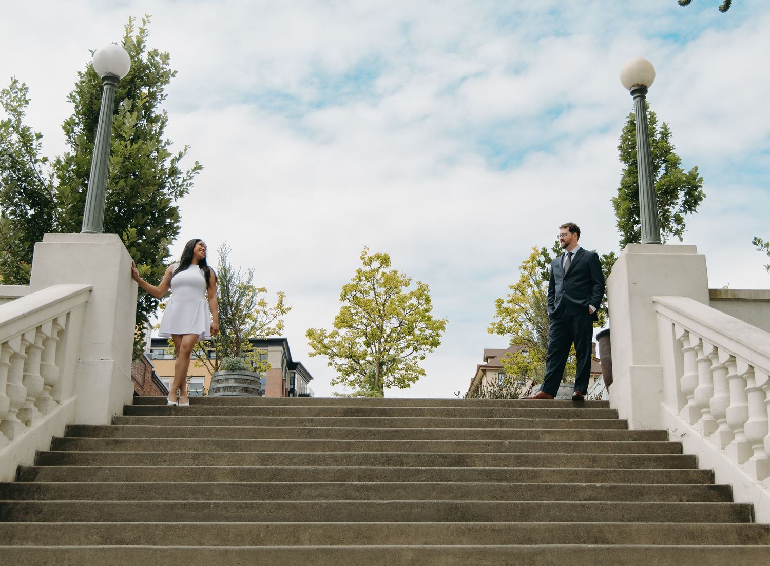 Couple standing apart on opposite sides of a grand stone staircase under a bright sky.