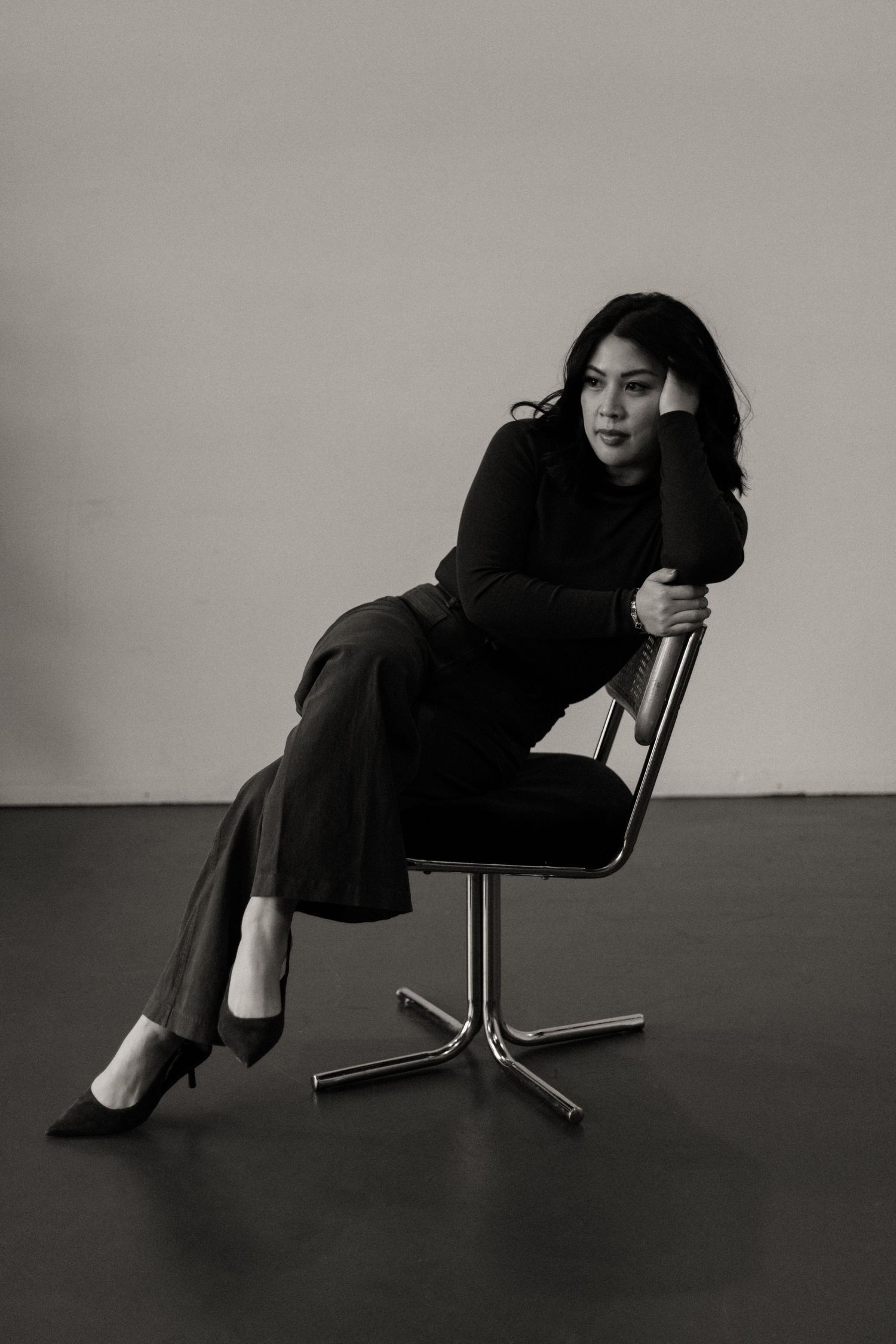Black and white studio portrait of woman in turtleneck sitting on chrome chair in relaxed pose.