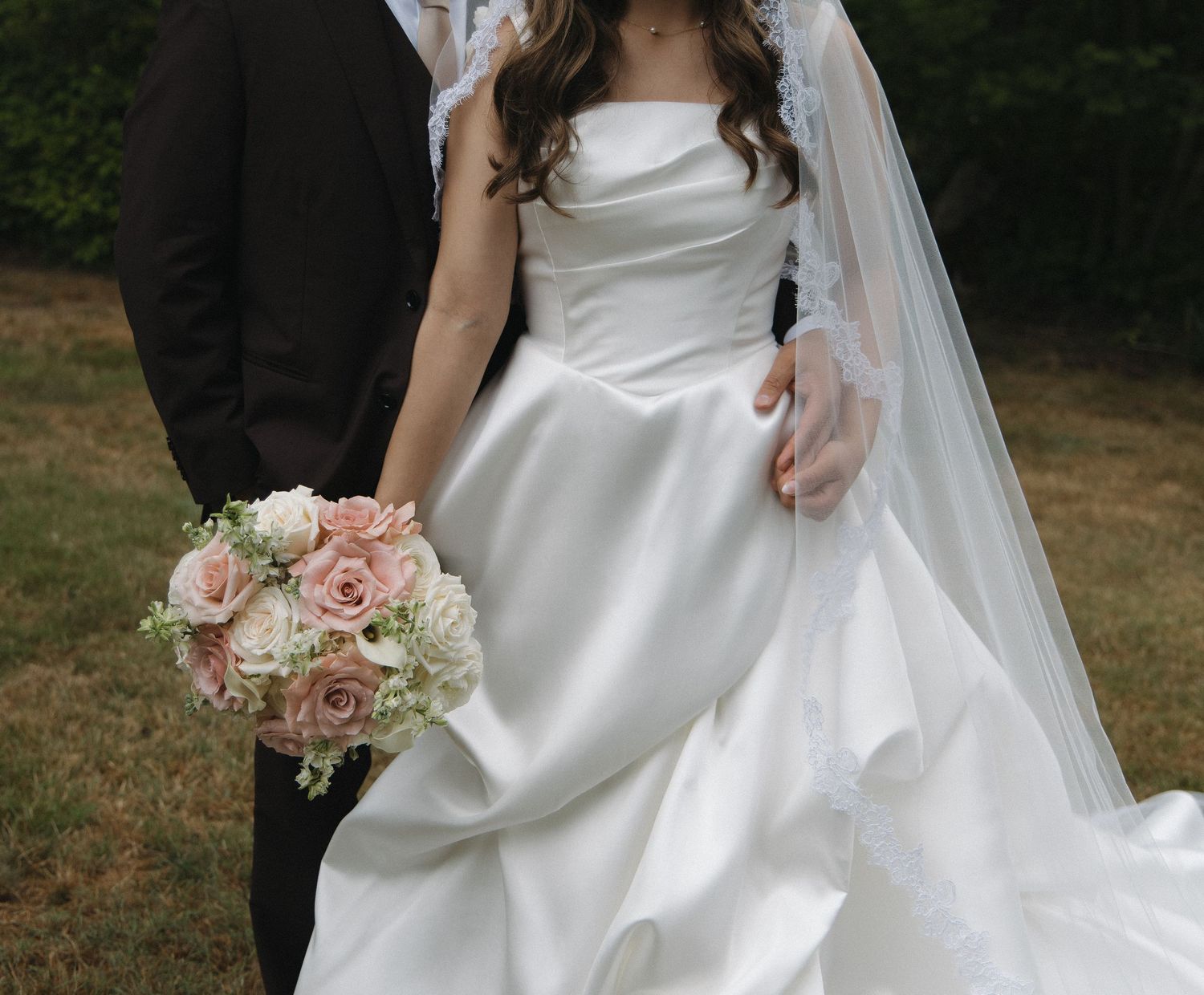 Bride in strapless white satin gown with lace veil holding pink rose bouquet beside groom outdoors.