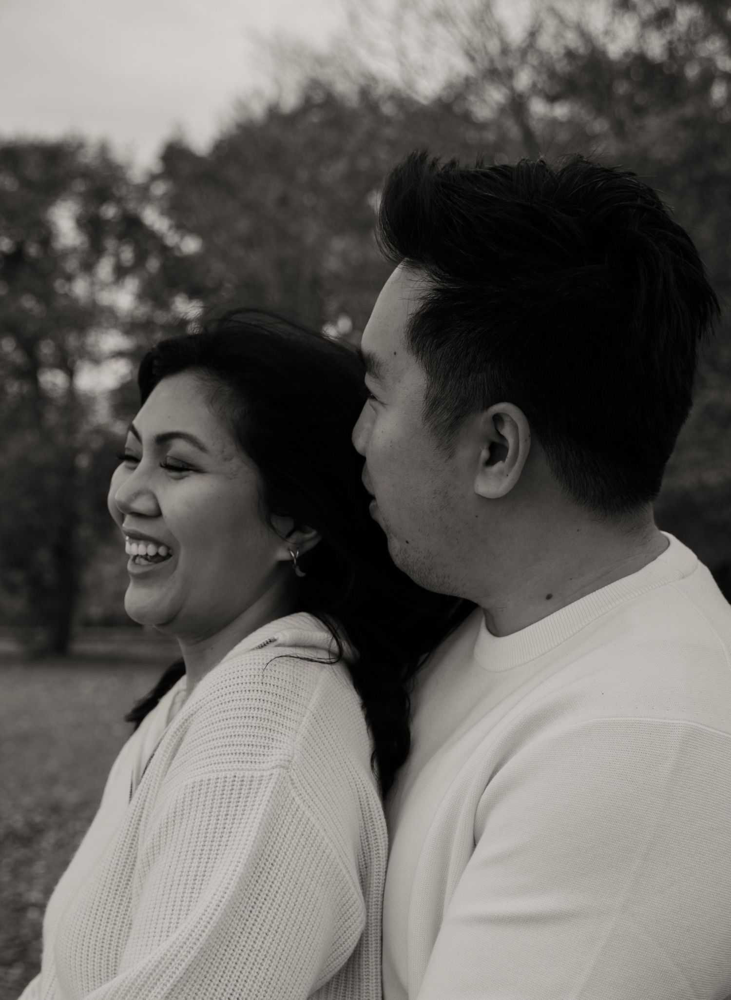 Black and white photo of a laughing woman and smiling man in a park, wearing white casual outfits.