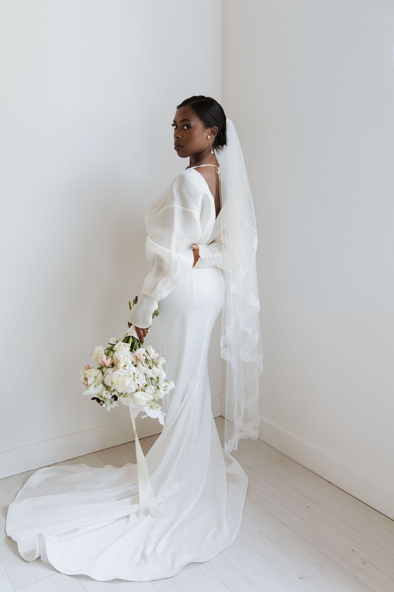 Elegant bride in a white gown with veil holding a bouquet of white flowers in a minimalist white room.