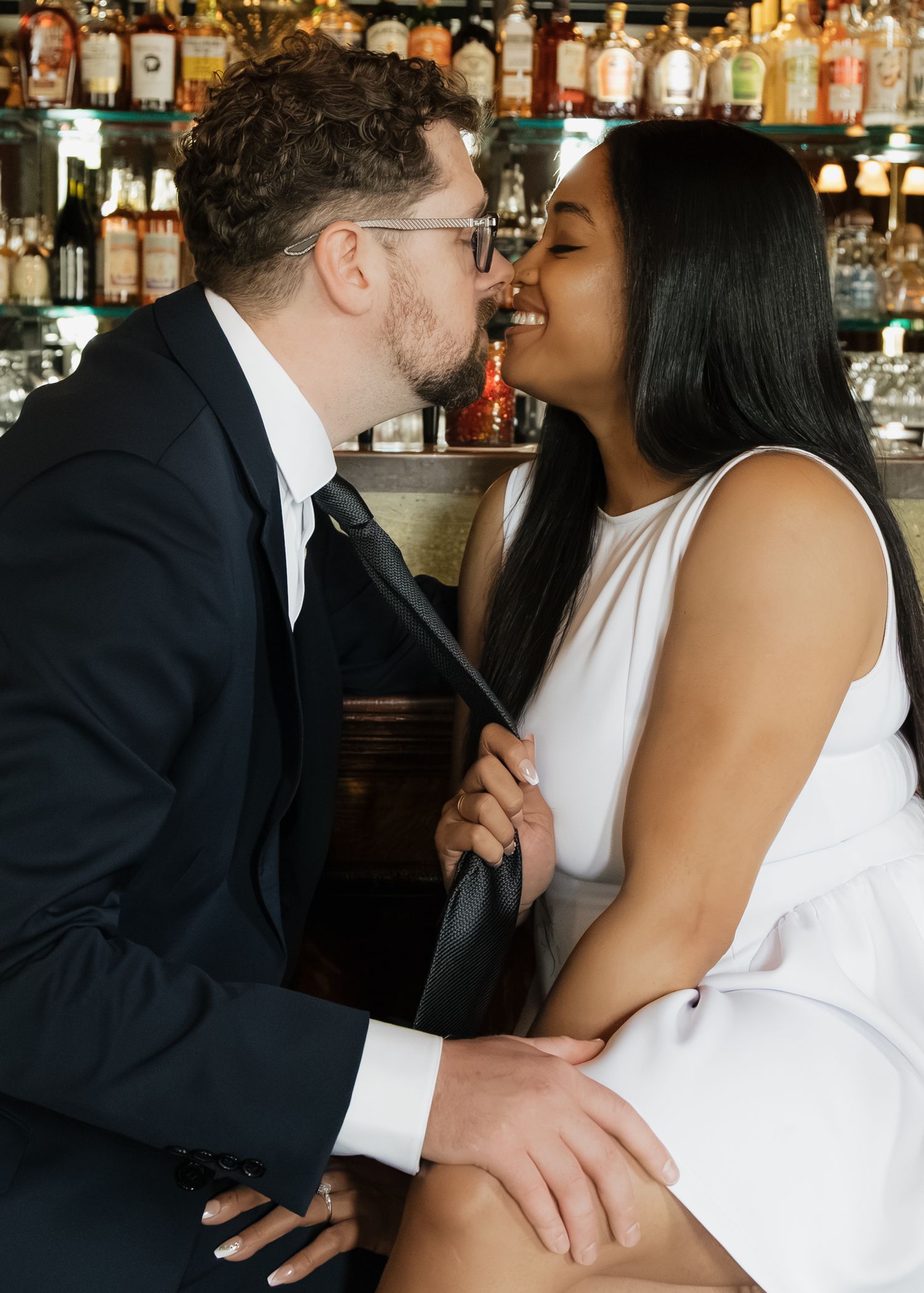 Close-up of interracial couple about to kiss, man in suit, woman in white dress at a bar.