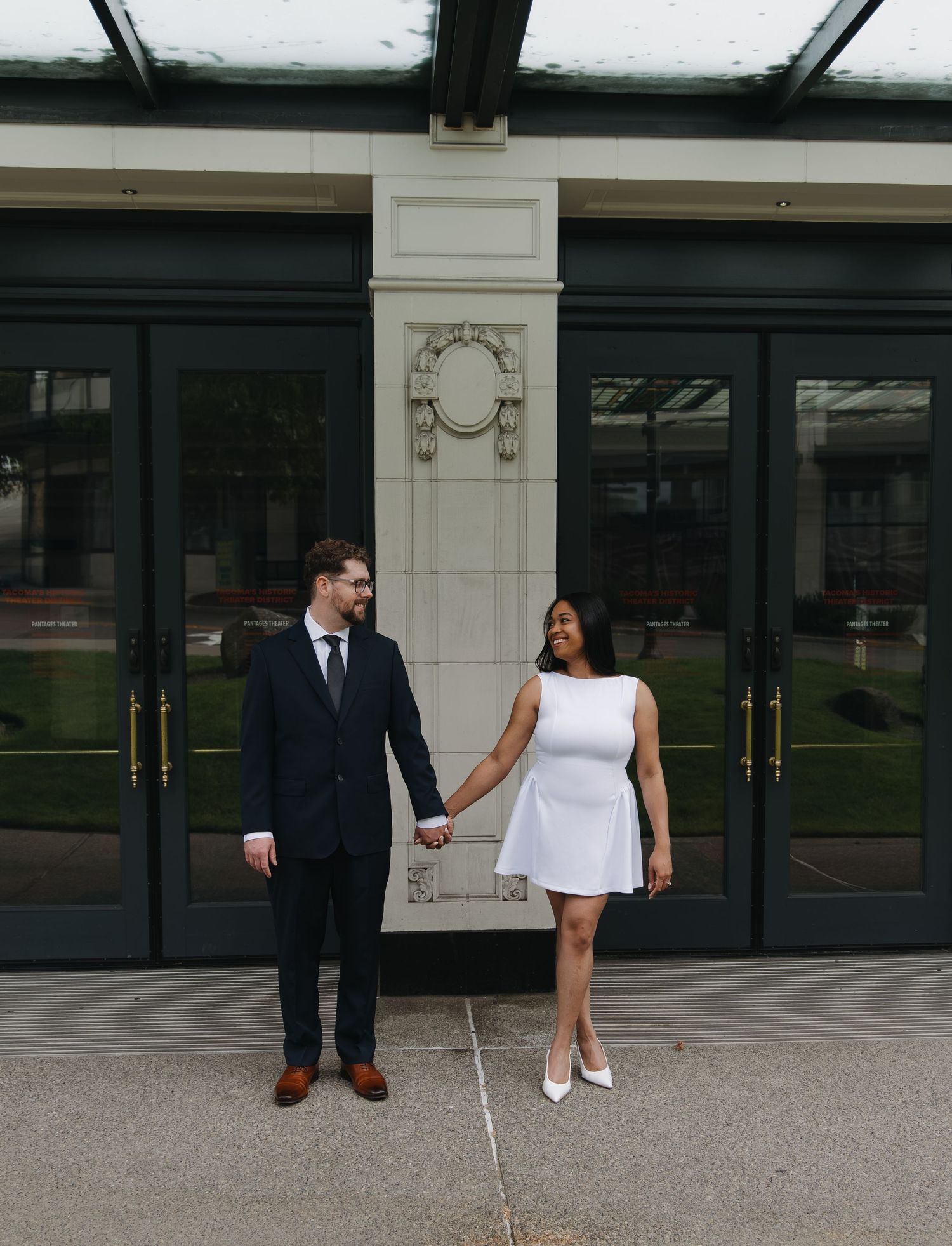 Interracial couple holding hands and gazing at each other outside elegant building, woman in white dress and man in dark suit.