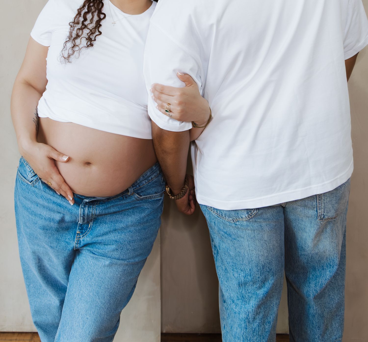 Pregnant couple in matching white tees and jeans cradle bare baby bump together.