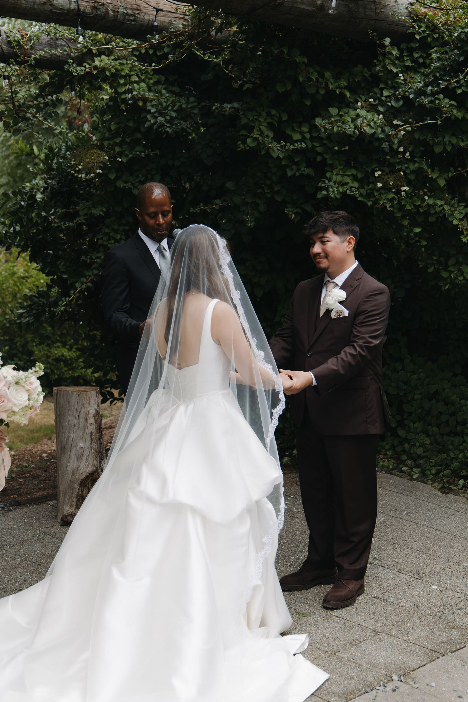 Bride and groom hold hands at outdoor wedding ceremony while officiant stands behind them.