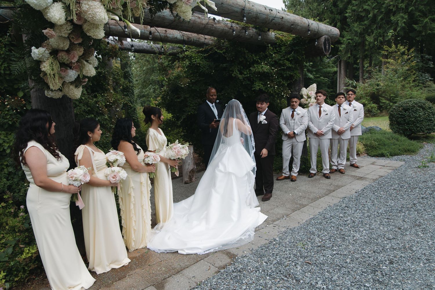 Wedding party stands at outdoor altar with bridesmaids in cream gowns and groomsmen in white suits.