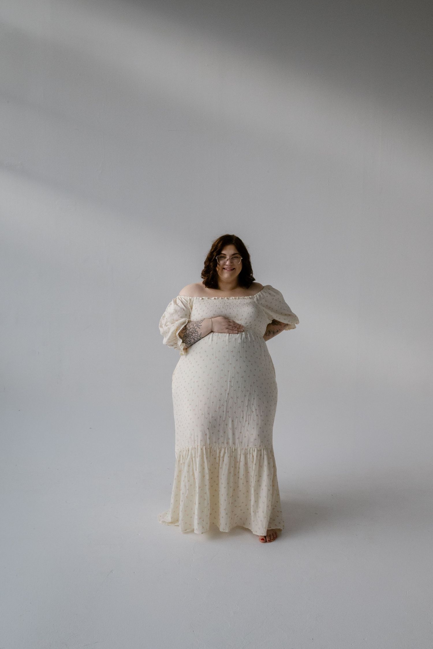 Pregnant woman in elegant white off-shoulder lace maternity gown poses in a bright studio.