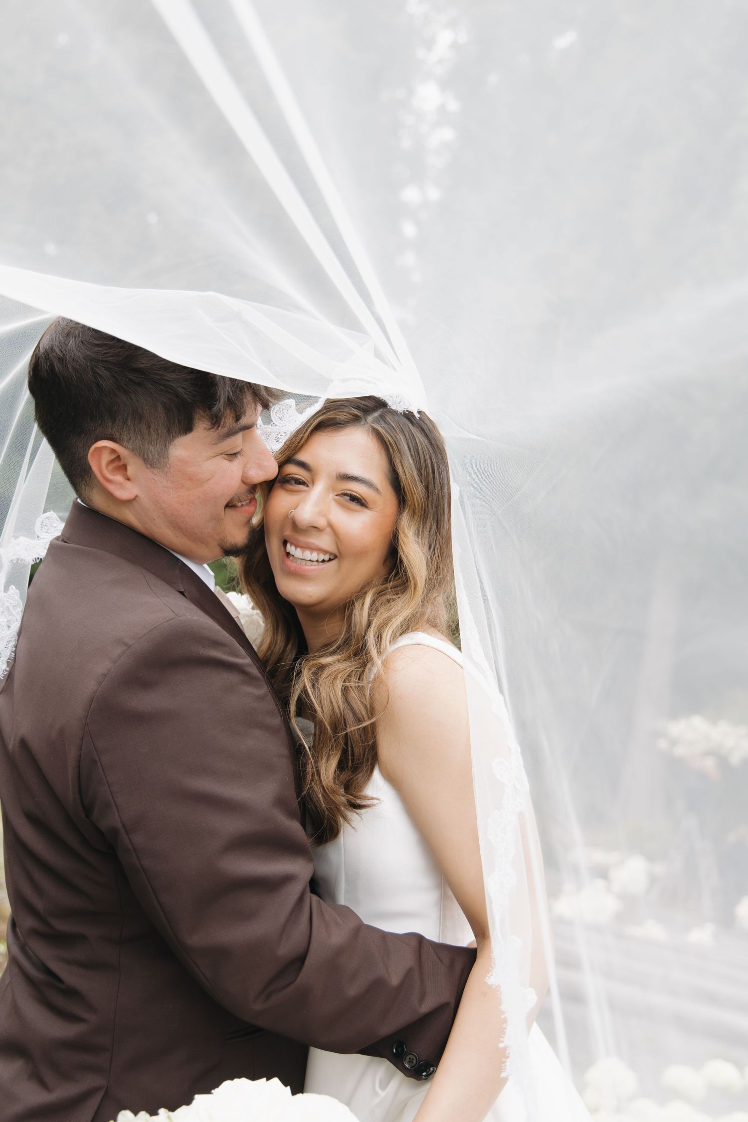 Bride and groom share a tender moment under a flowing white veil, bride smiling joyfully at outdoor wedding.