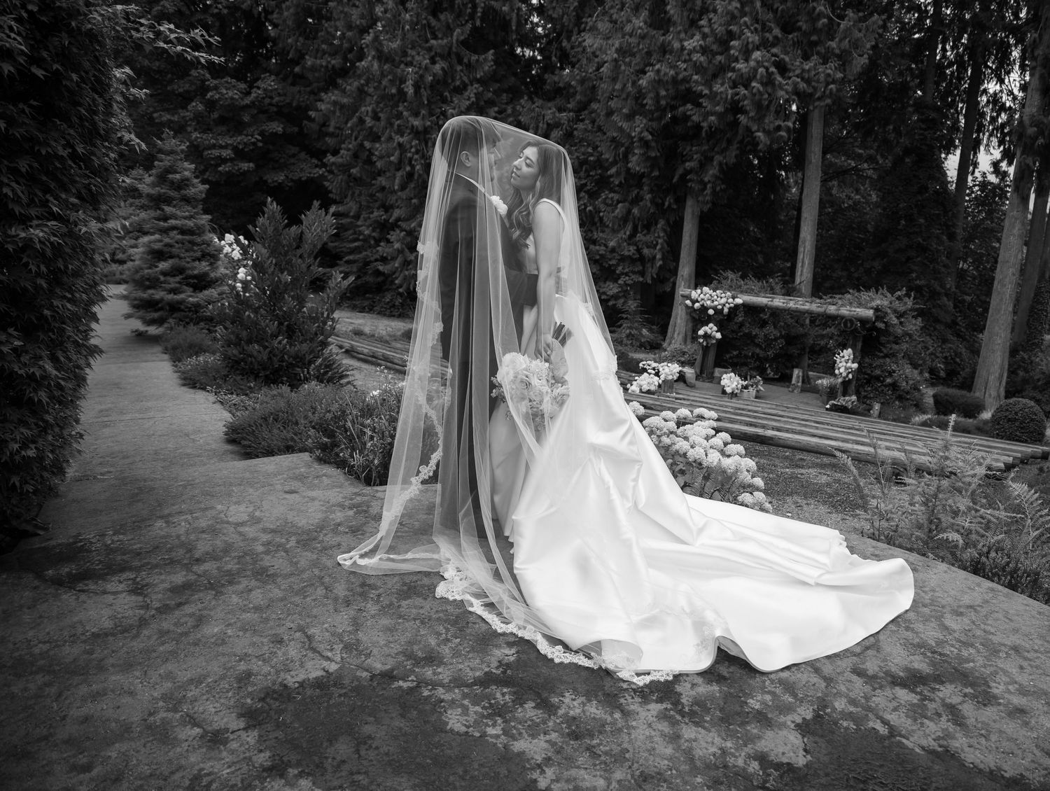 Black and white photo of a bride and groom kissing under a long veil in a garden setting.