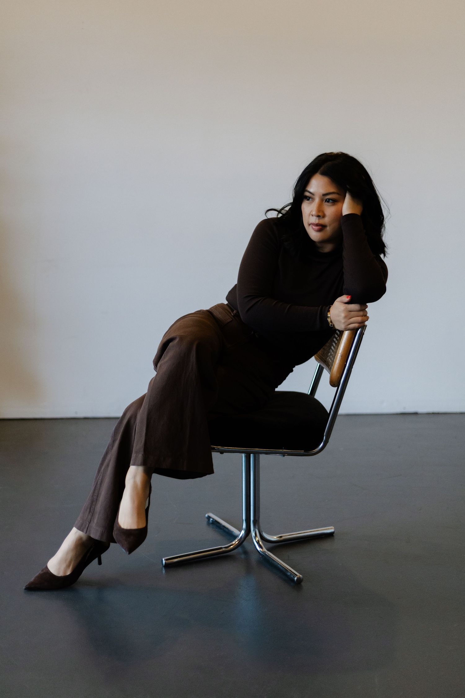 Woman in dark turtleneck and wide-leg pants sits sideways on chrome chair in moody studio portrait.