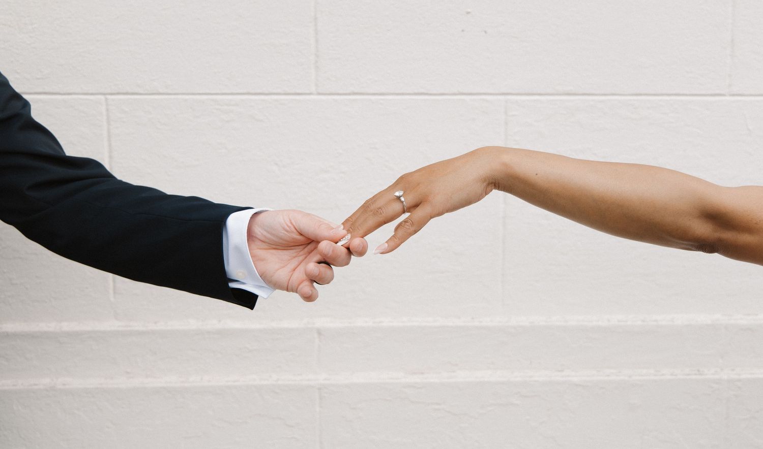 Close-up of interracial couple holding hands, bride wearing diamond engagement ring against white wall.
