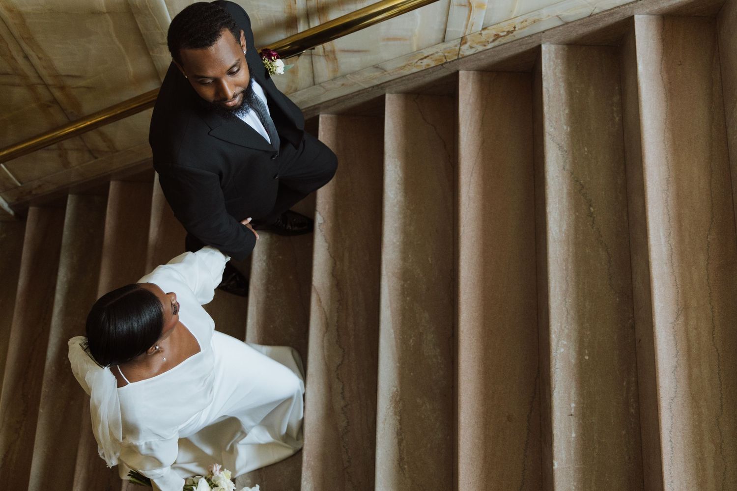 Aerial view of bride and groom in wedding attire standing on elegant marble staircase with tall columns.