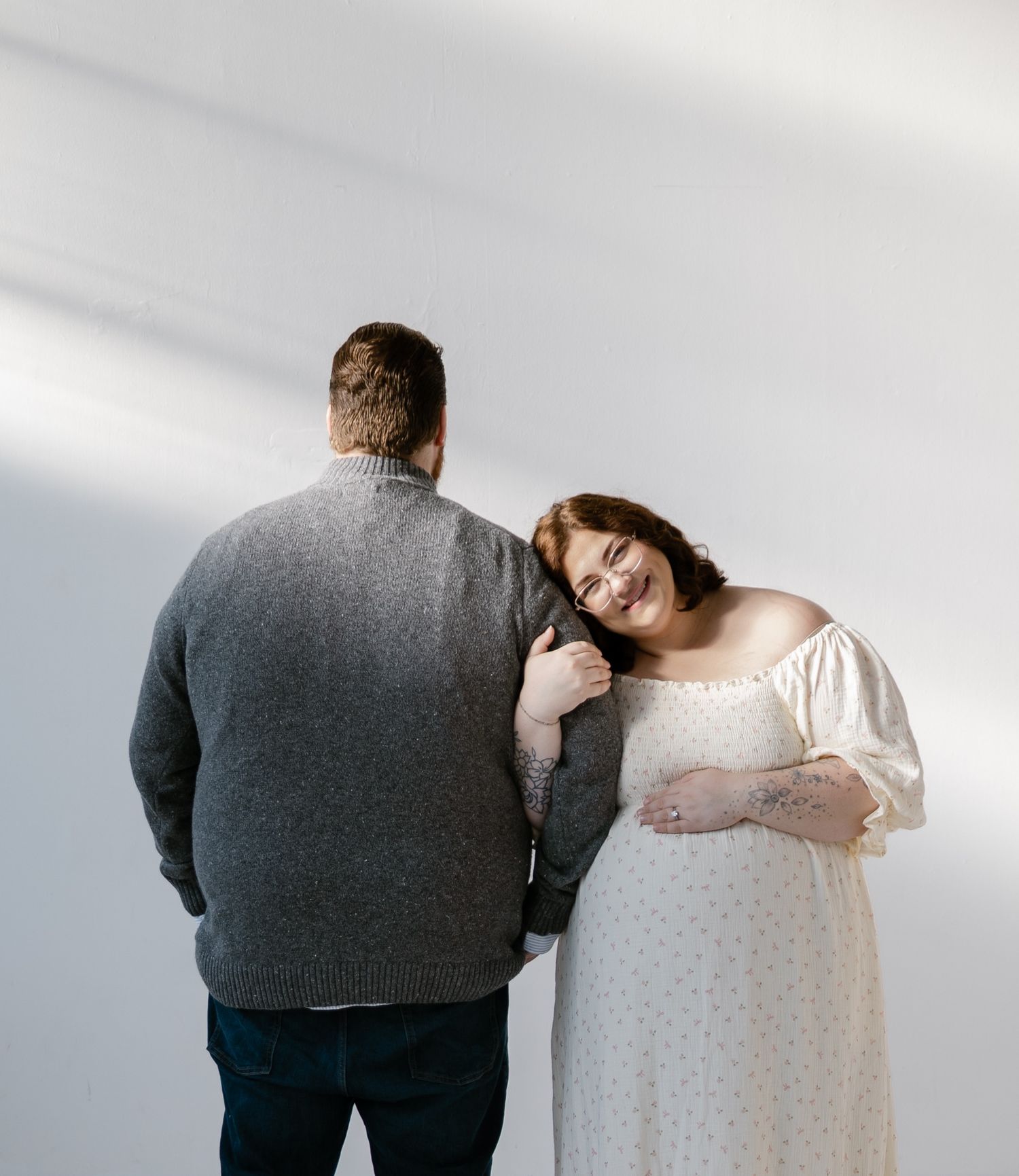Smiling pregnant woman in white floral dress leans on partners shoulder in bright studio.