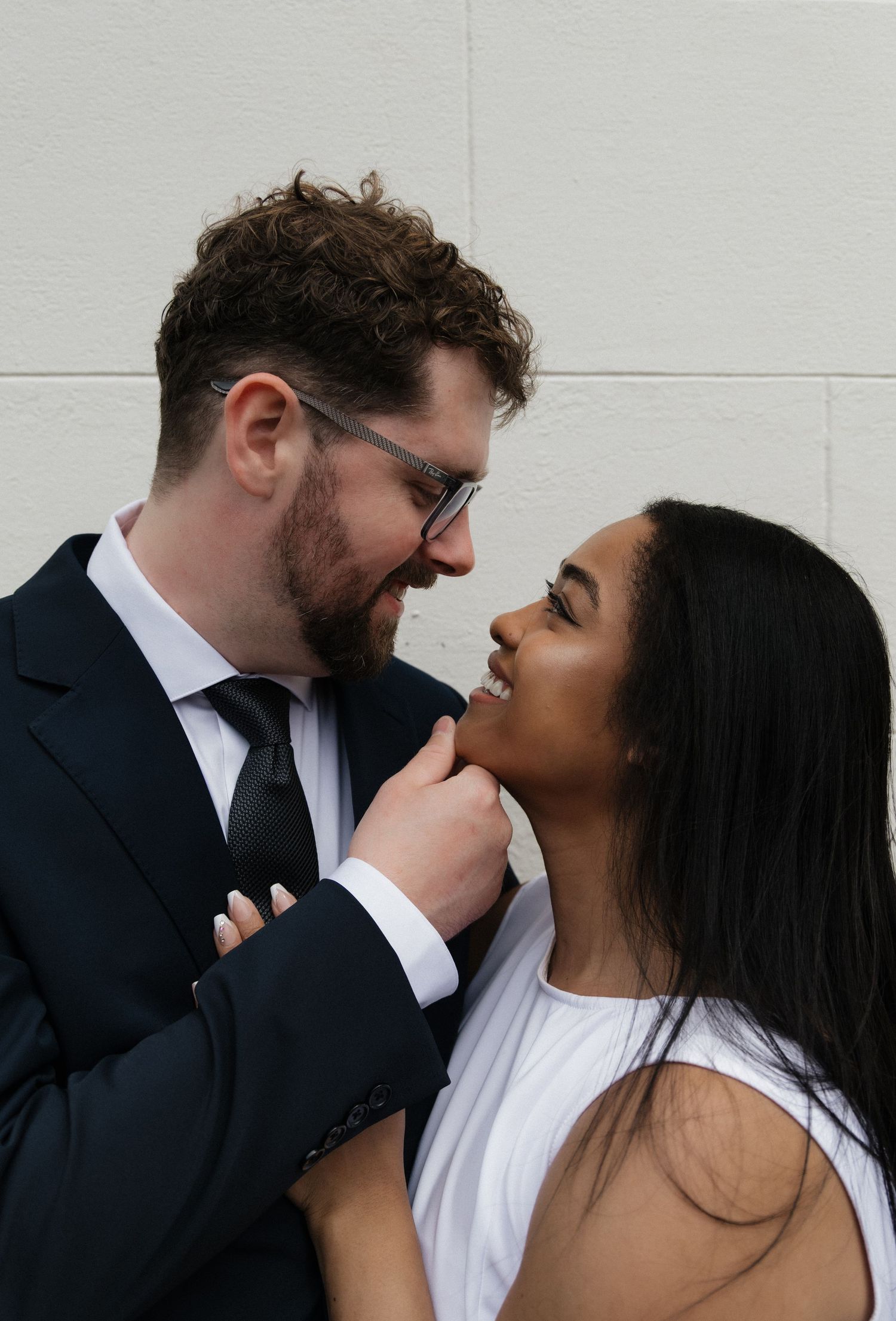 Interracial couple sharing a tender moment, groom in navy suit gently touching bride's chin outdoors.