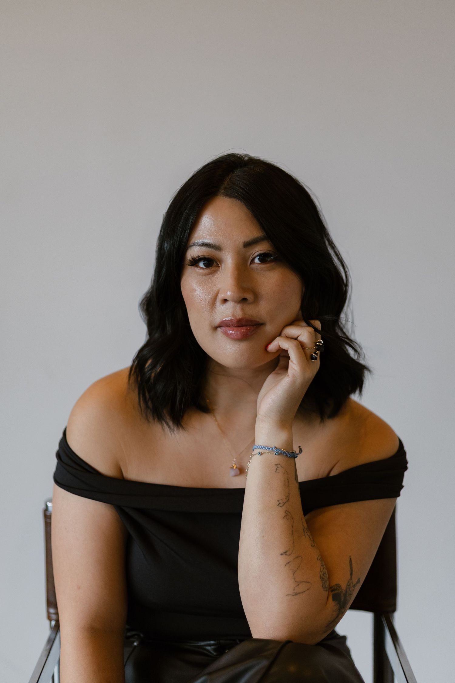 Woman in black off-shoulder top sits on chair with hand near face, showcasing tattoo and jewelry.