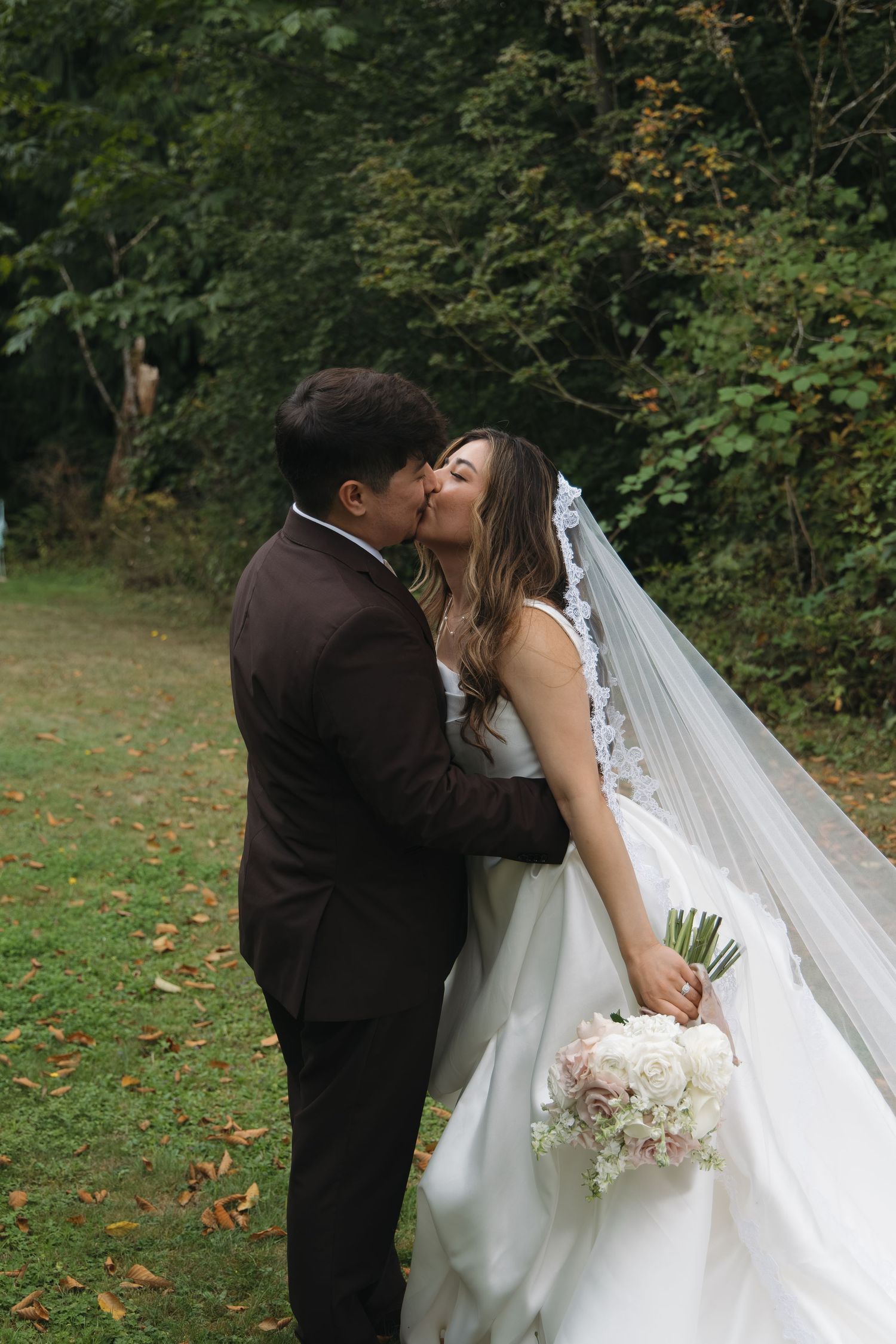 Bride and groom kissing outdoors, bride in white gown with lace veil holding pastel bouquet.