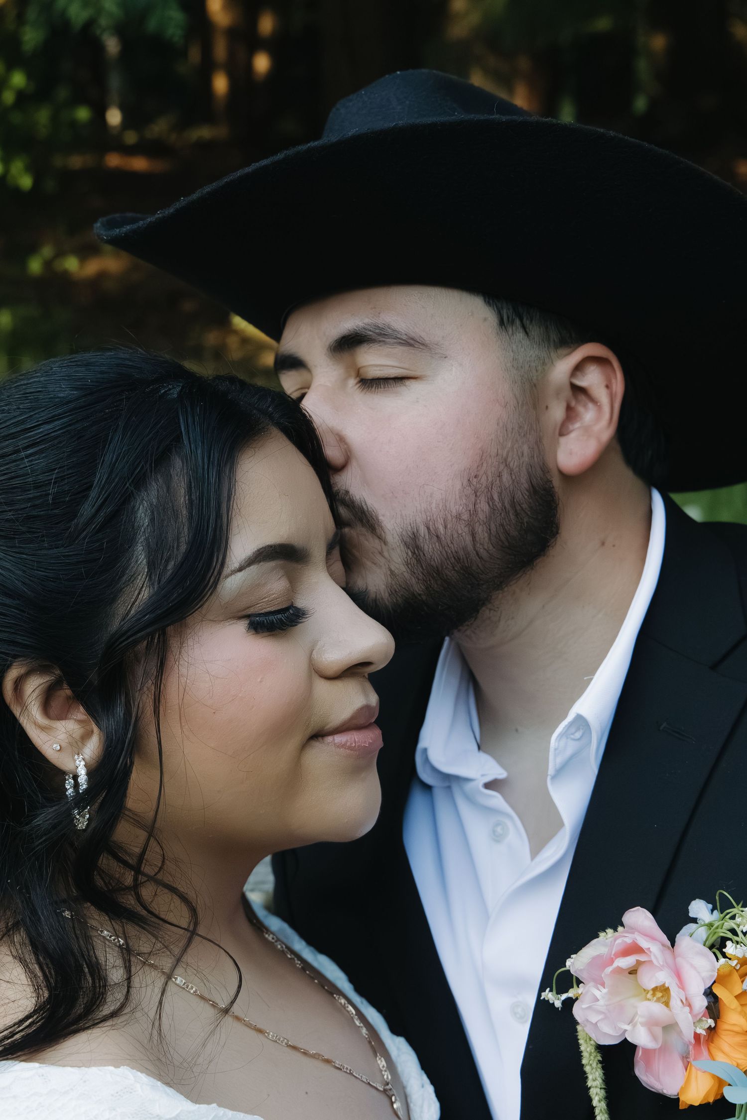 Groom in black cowboy hat tenderly kissing bride on cheek in romantic outdoor wedding portrait.