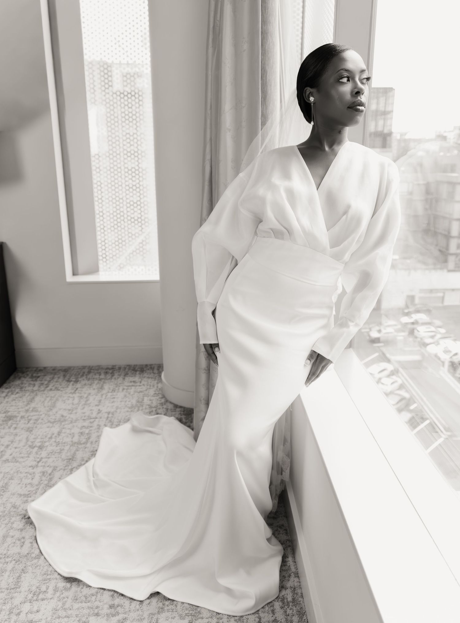 Stunning black-and-white portrait of bride in dramatic white gown with train and veil gazing out hotel window.