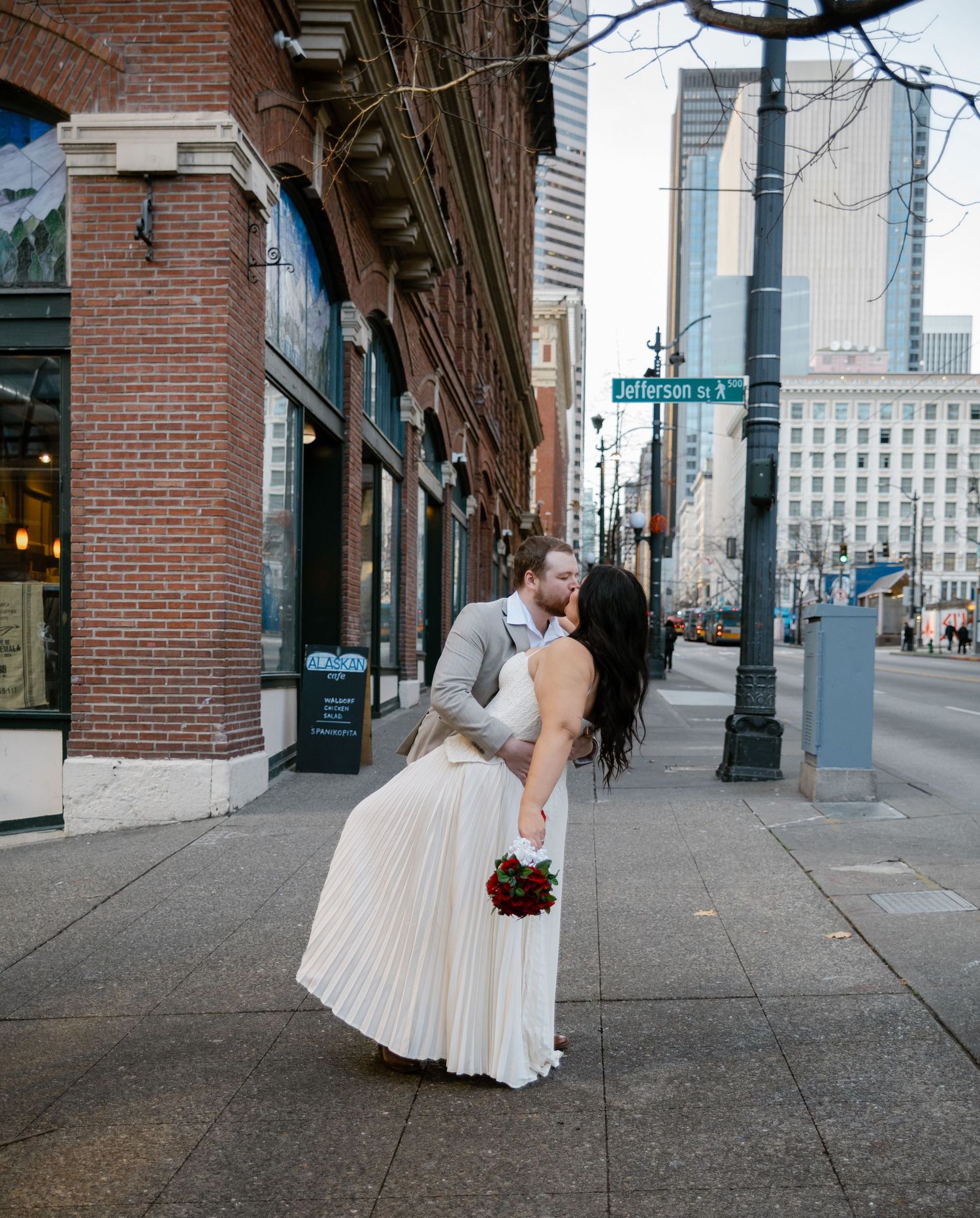 Bride and groom sharing a romantic dip kiss on a city sidewalk near Jefferson St, bride holding red roses.