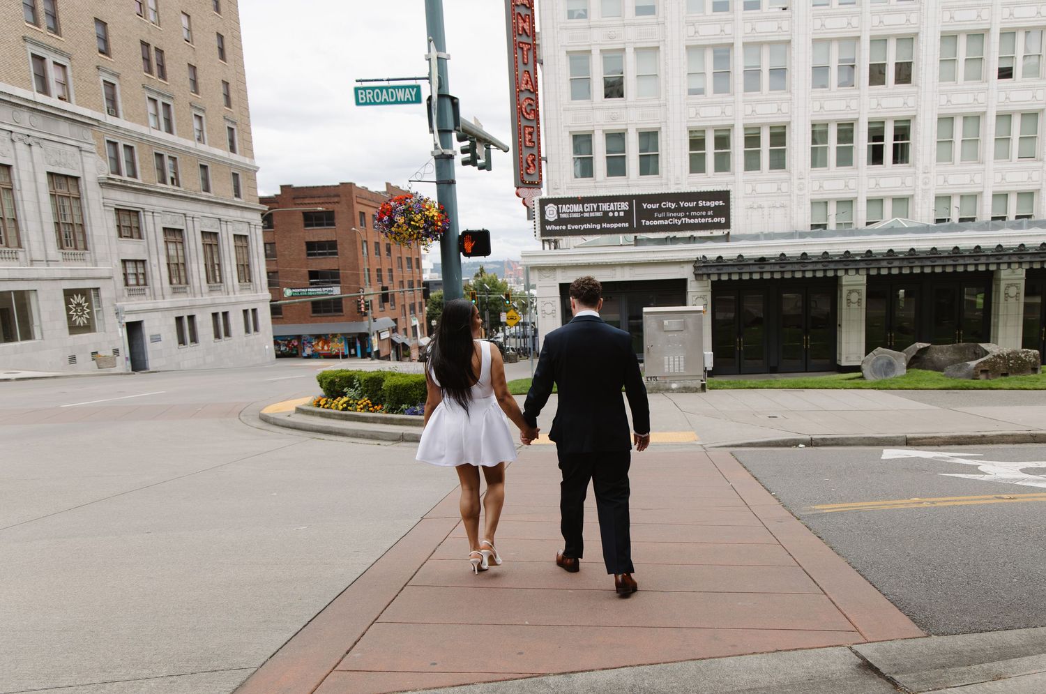 Couple walking hand in hand down a city sidewalk, woman in white dress and man in black suit.