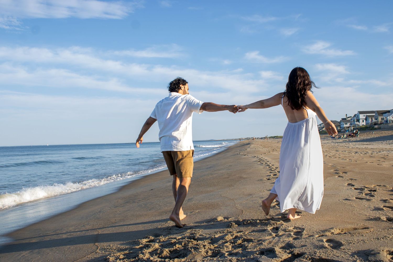 A couple walks hand in hand along a sunny beach near the ocean waves.