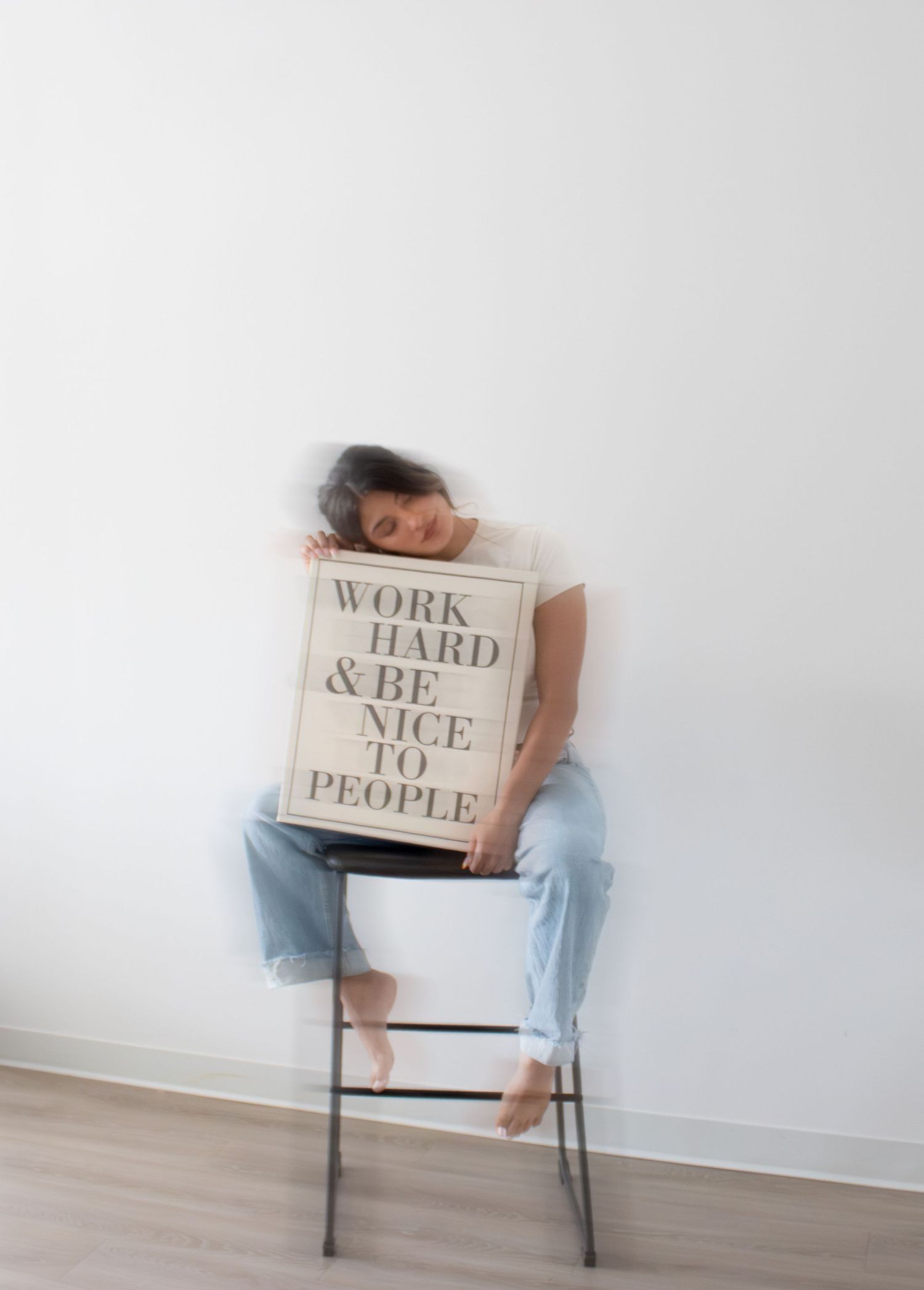 Person sitting on a chair holding a motivational sign reading Work Hard & Be Nice To People against a white wall.