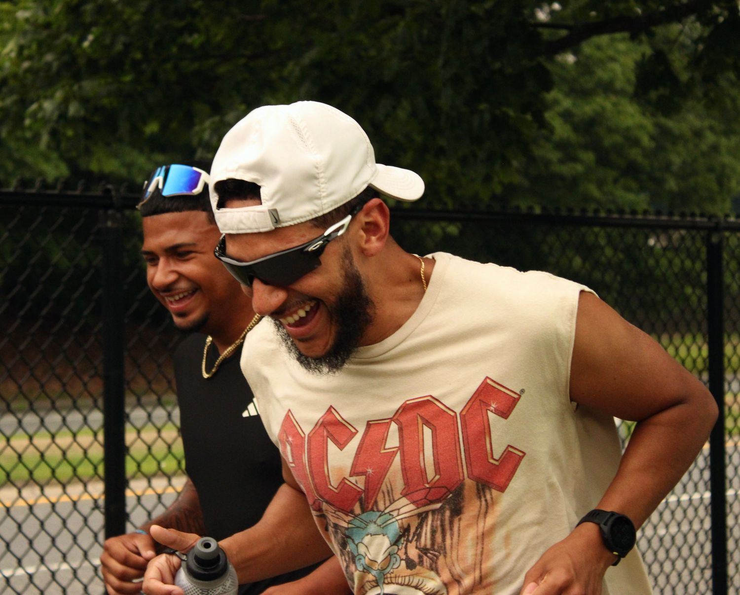 Two men laughing outdoors, one wearing an AC/DC shirt and white cap, near a chain-link fence.